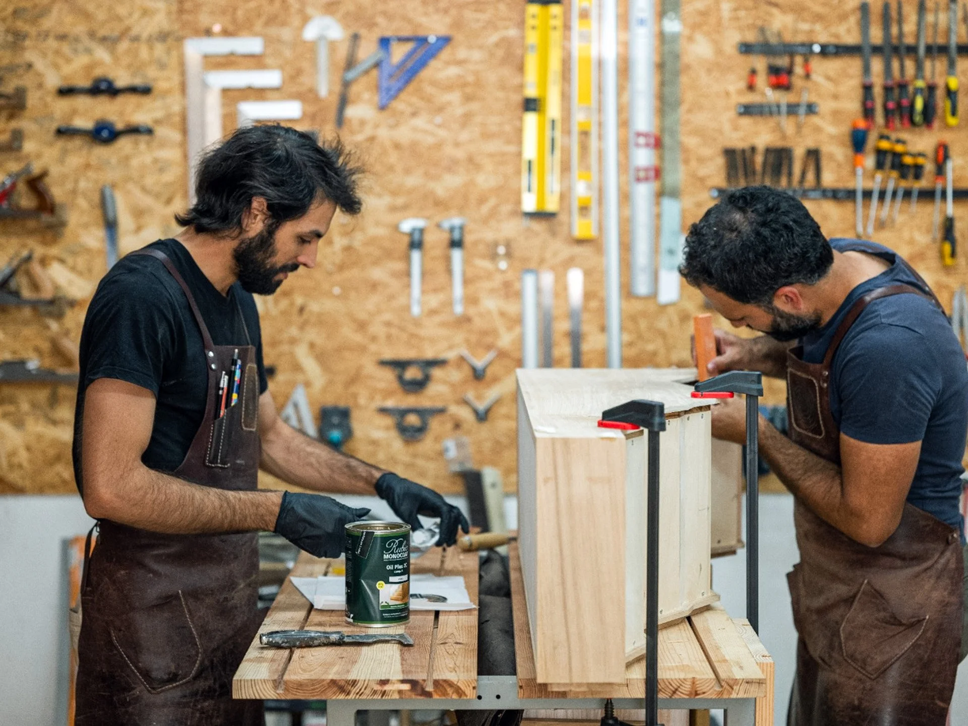 Manel a envernizar uma peça de madeira e Zé a fazer uma colagem no atelier Nó, com várias ferramentas na parede ao fundo.