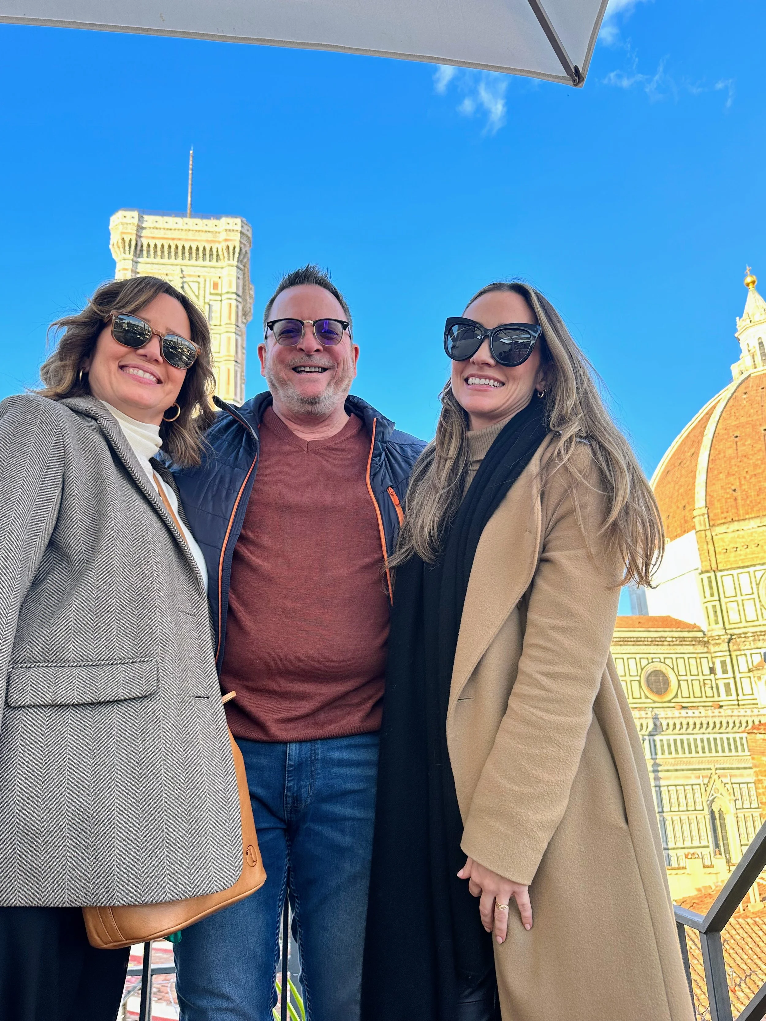 Three people smiling outdoors with a historic European building featuring a dome and a tower in the background, under a blue sky.