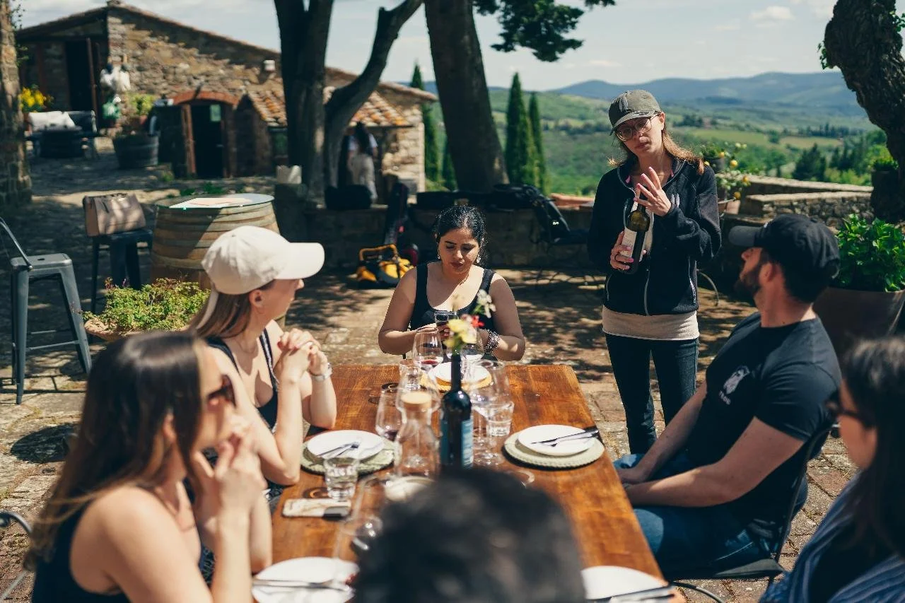 Group of people having an outdoor meal at a rustic vineyard or countryside setting with scenic hills in the background, with a woman standing and talking while others listen at a wooden table.