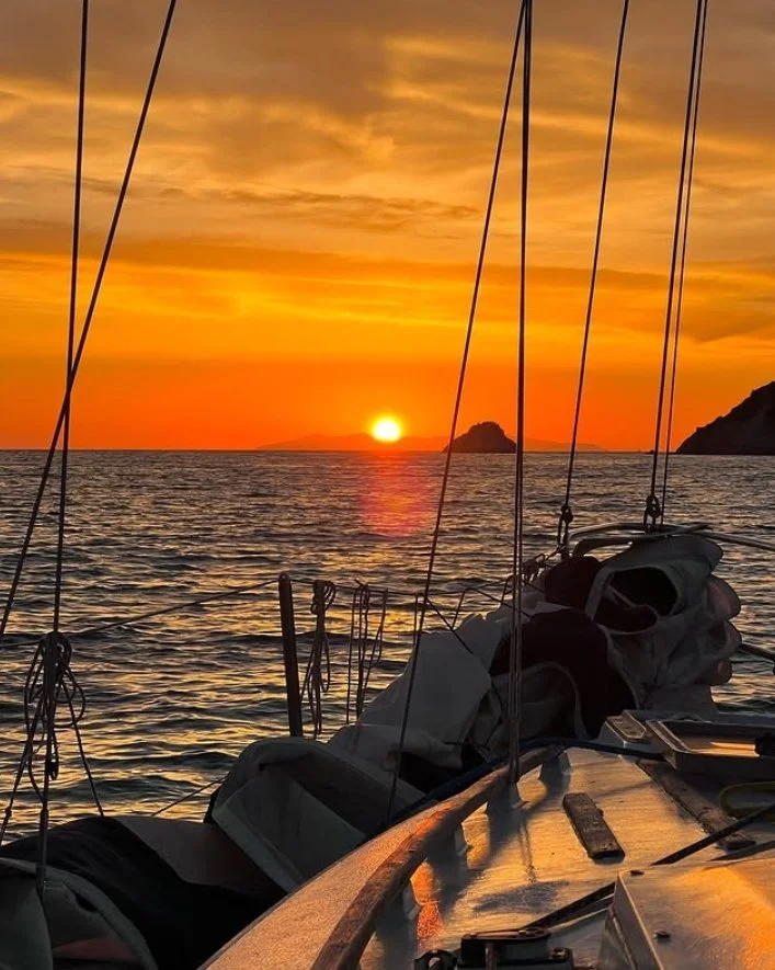 View of a sunset over the ocean as seen from a sailboat with ropes and equipment on deck, and small islands in the distance.