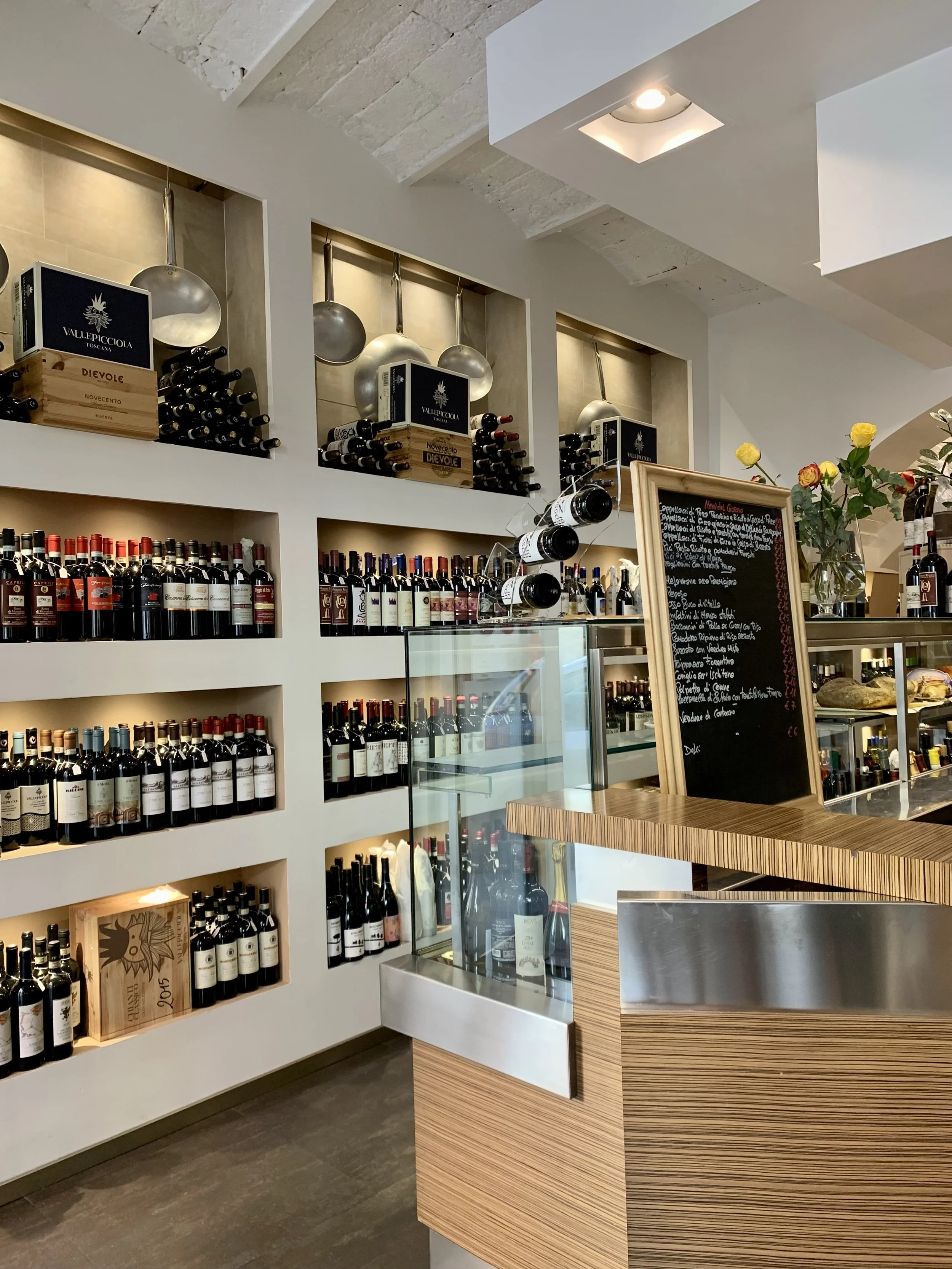 Interior of a wine shop or restaurant with shelves filled with bottles of wine, wine boxes on the upper shelves, a chalkboard menu, and a glass display case.