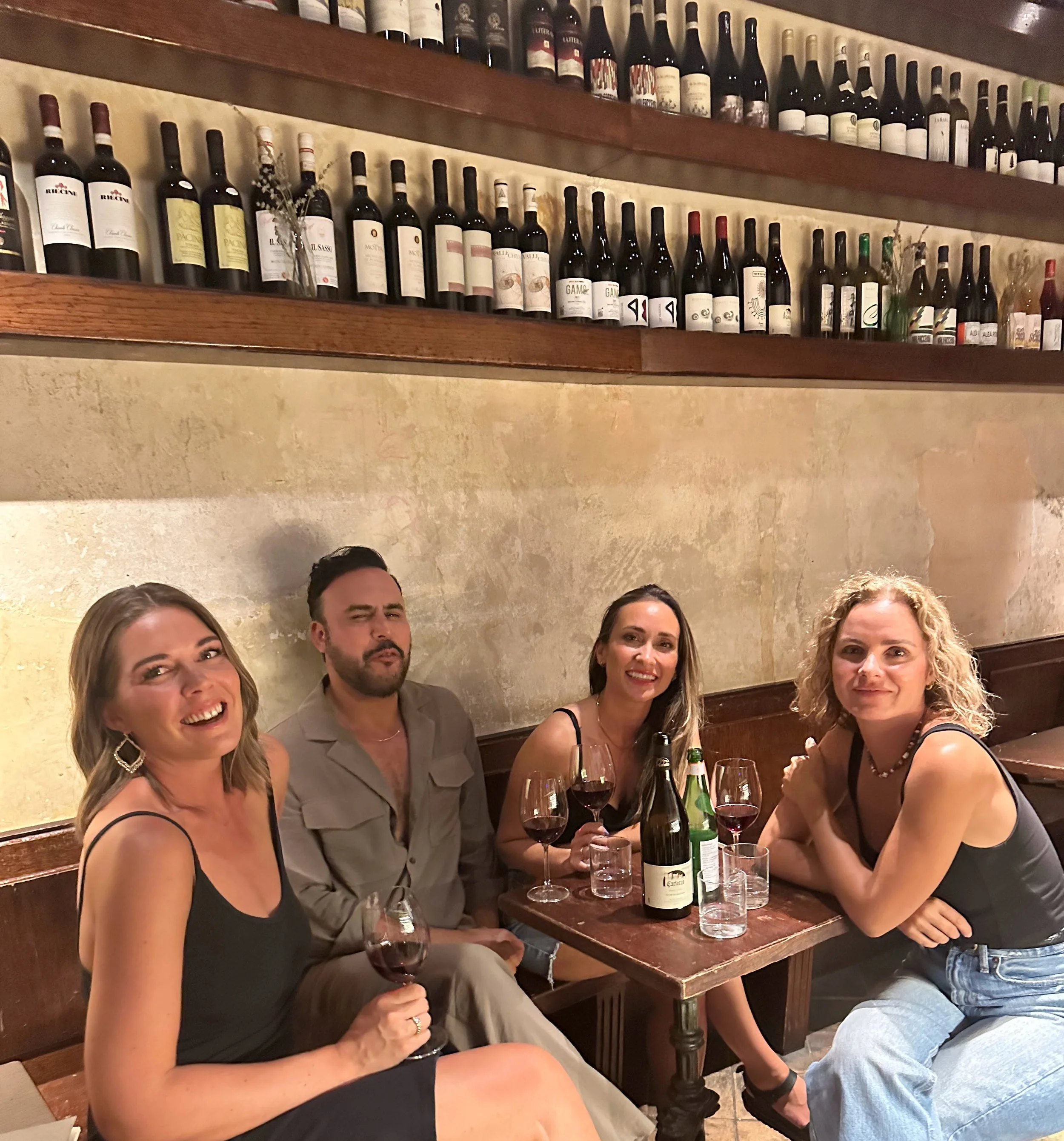 Four friends sitting at a wooden table in a cozy restaurant, enjoying glasses of red wine, with a wall lined with wine bottles behind them.