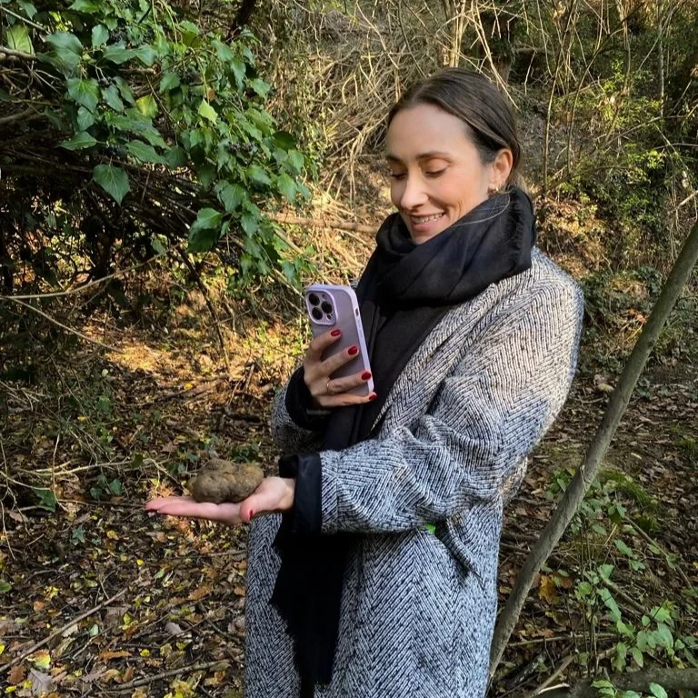A woman with dark hair, wearing a gray coat and black scarf, stands outdoors in a wooded area, smiling while looking at a small rock she is holding in her palm. She is taking a selfie with her phone.