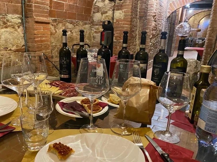 A dining table set with empty wine glasses, a plate with a small piece of food, and a tasting platter of meats and cheeses. Behind the table, there are five bottles of red wine, a paper bag, and other bottles of beverages. The background features a r