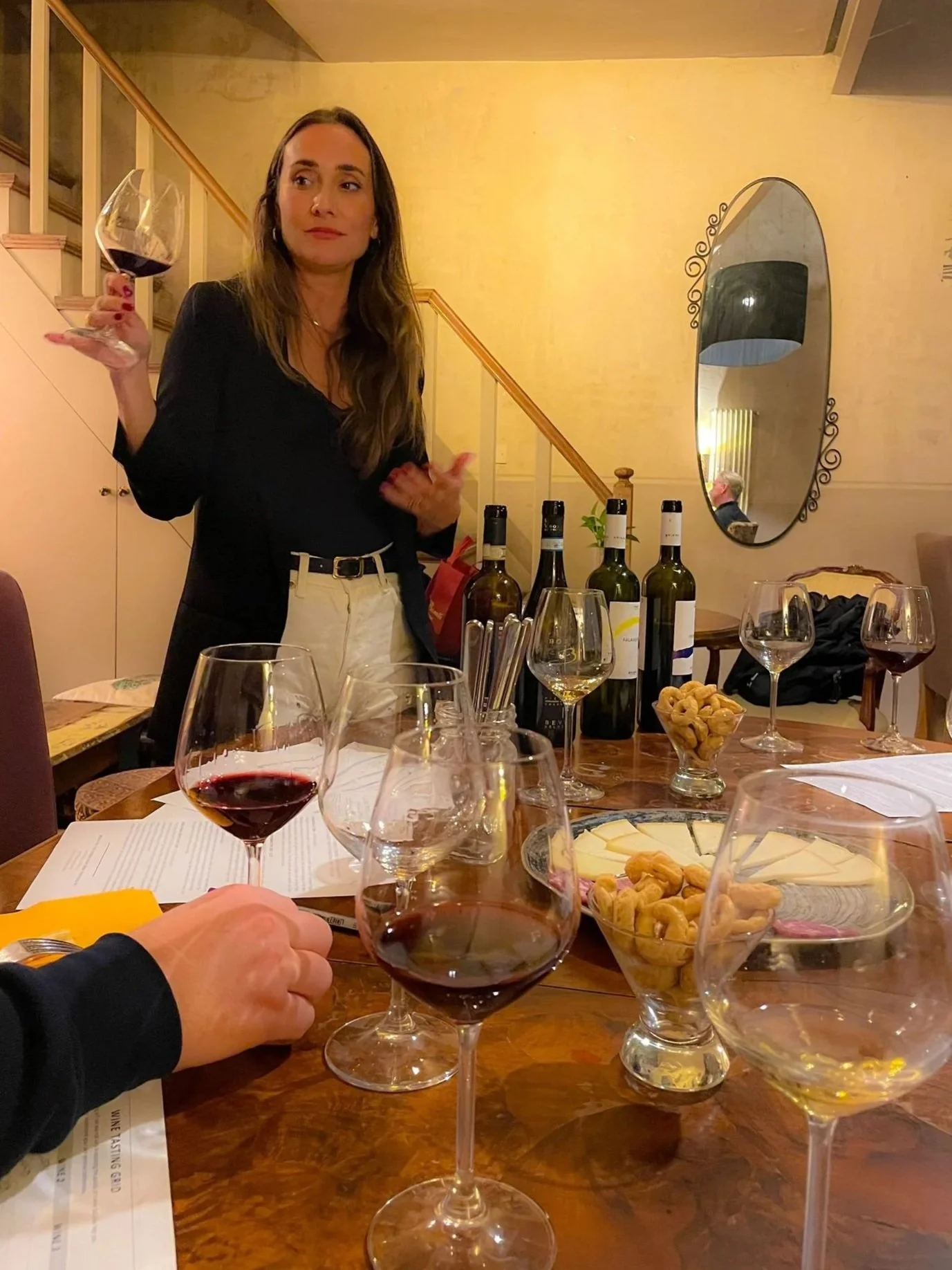A woman standing at a table with various wine glasses, wine bottles, cheese, and nuts, holding a glass of red wine, in a cozy room with a yellow wall and a large oval mirror.