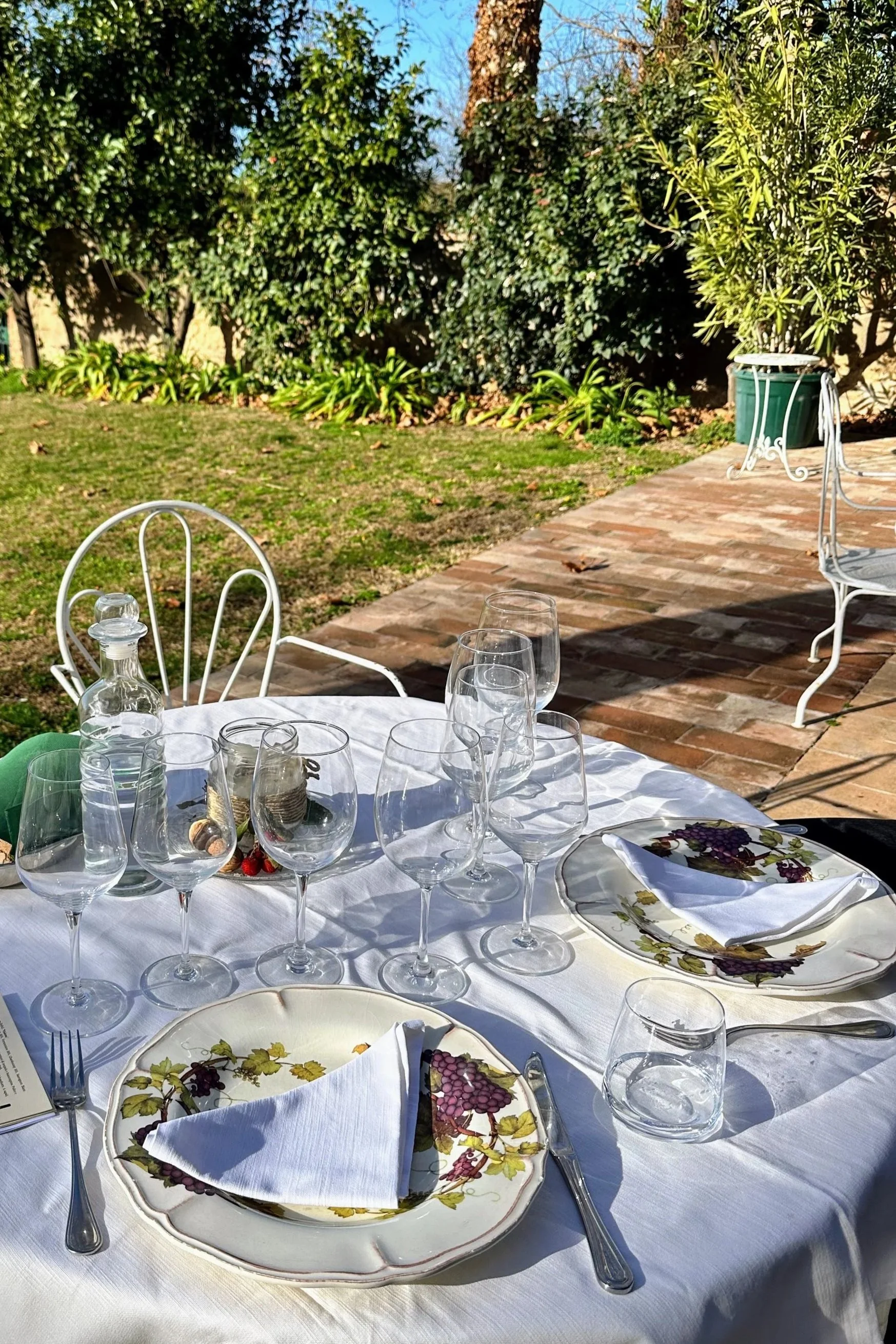 An outdoor dining table set with plates, glasses, and napkins on a white tablecloth, with a garden and patio in the background.