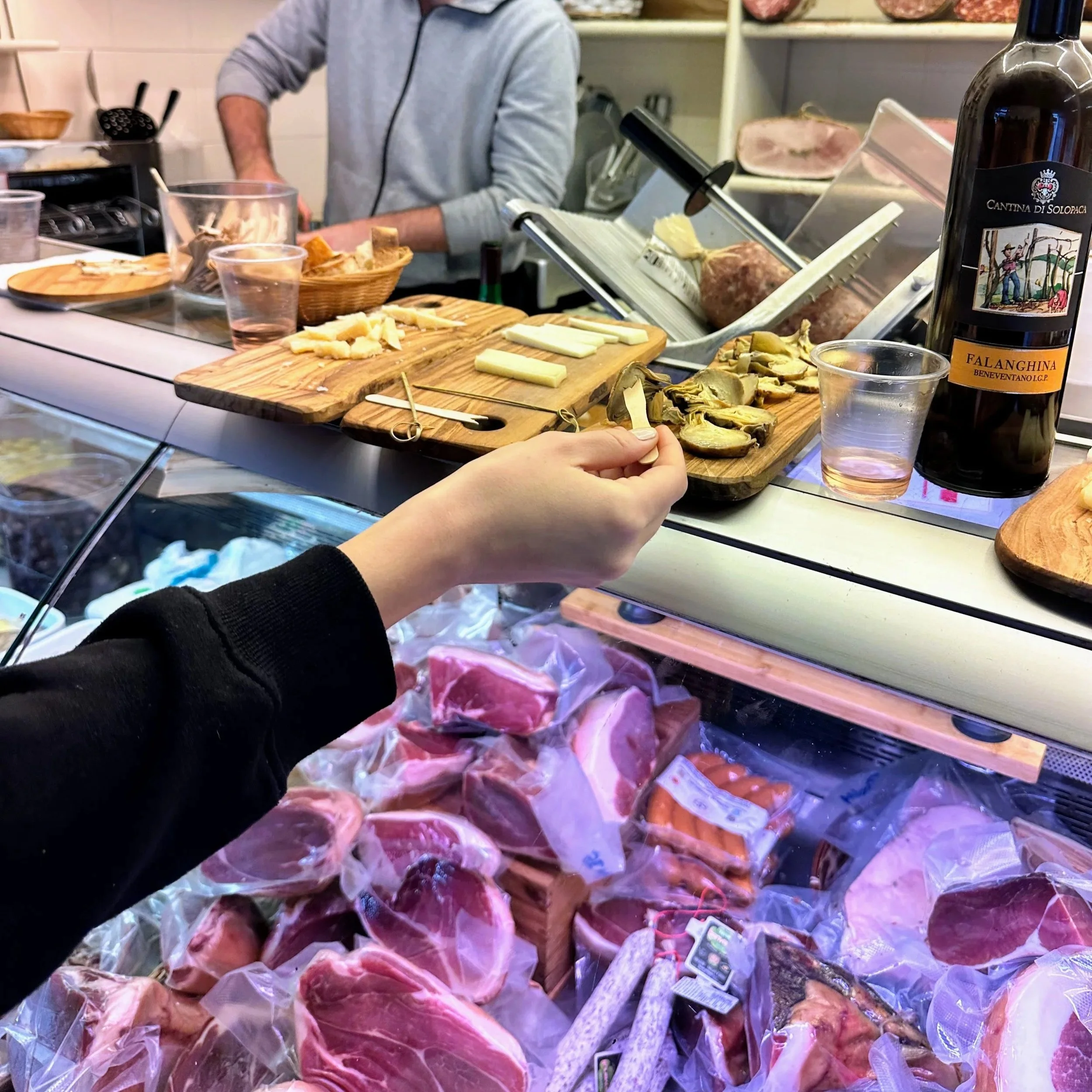 A person holding a slice of cheese in a deli or grocery store, with various meats, cheeses, and wine bottles on the counter and shelves.