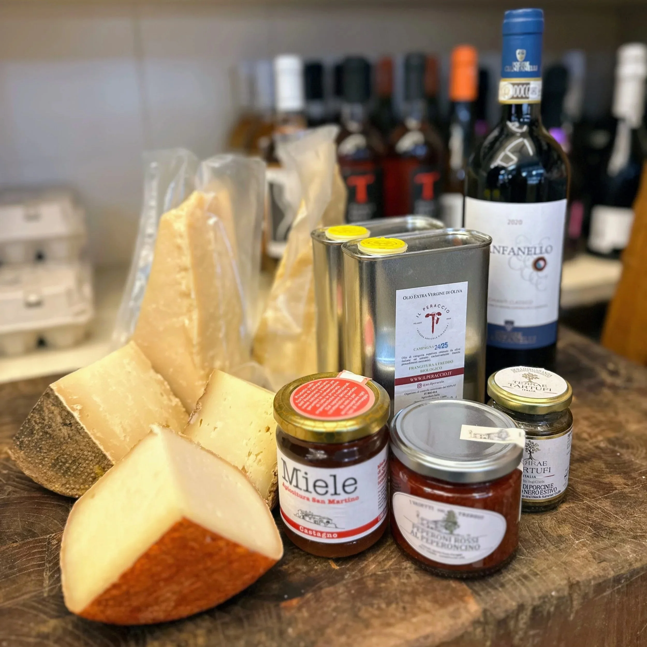 A variety of cheeses, jars of jam, a bottle of red wine, a tin of extra virgin olive oil, and other Italian food items on a wooden surface.