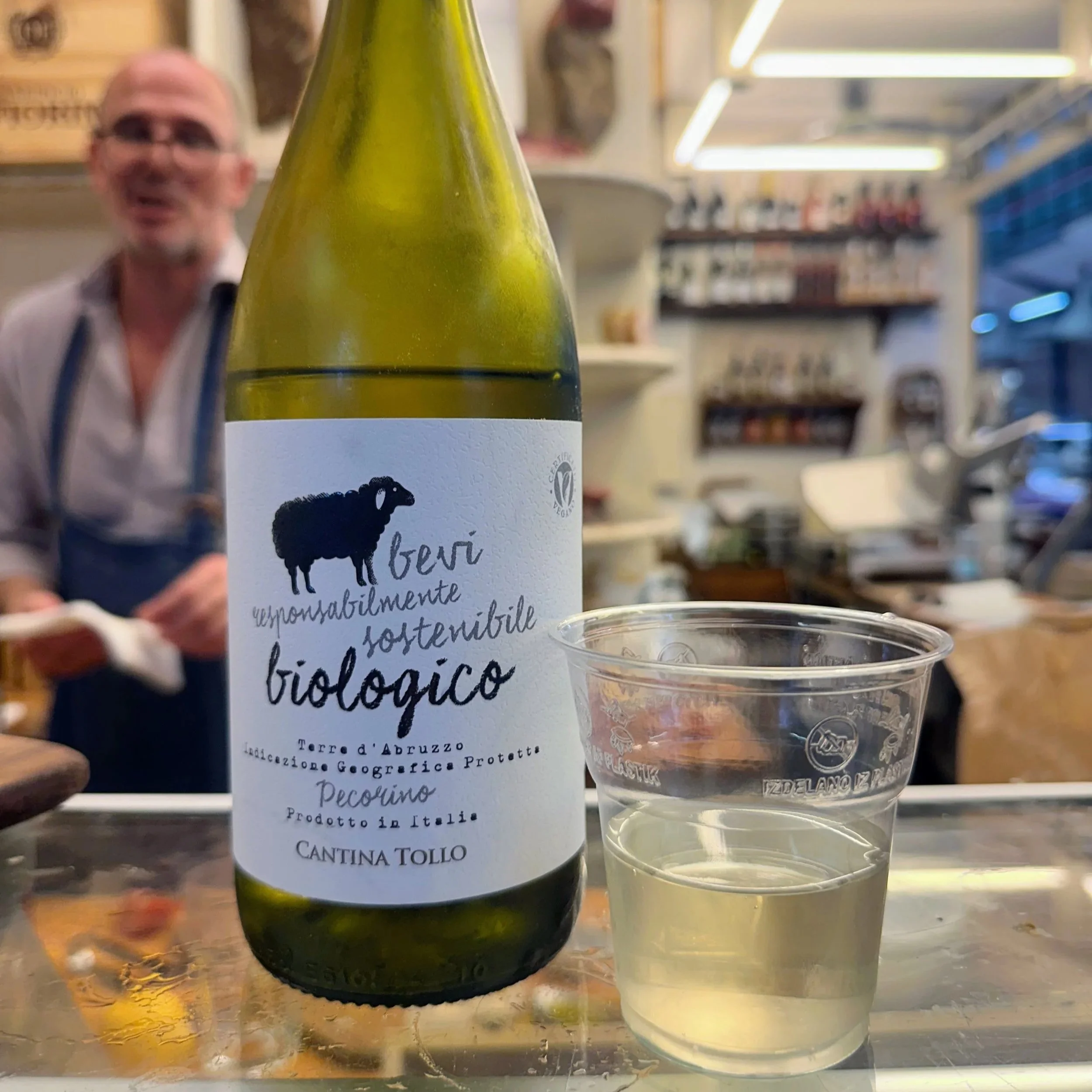 A bottle of organic Italian wine, Pecorino, from Cantina Tollo, placed on a counter with a clear plastic cup of white wine next to it. In the background, a man wearing glasses and an apron is smiling.