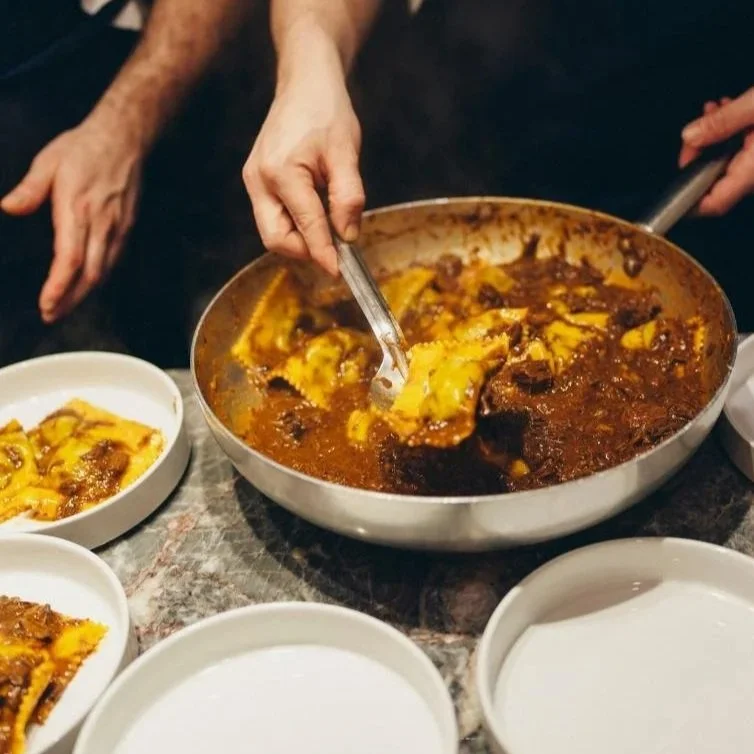 Person serving near a large pan of yellow and brown stew or curry, with additional empty bowls on a table.