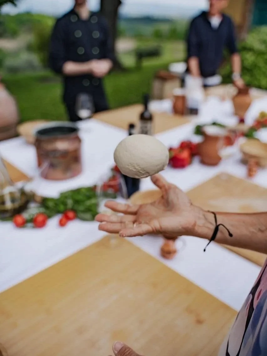 A person holding a white ice cream ball on a cone, with a blurred outdoor setting and a table with various food items in the background.