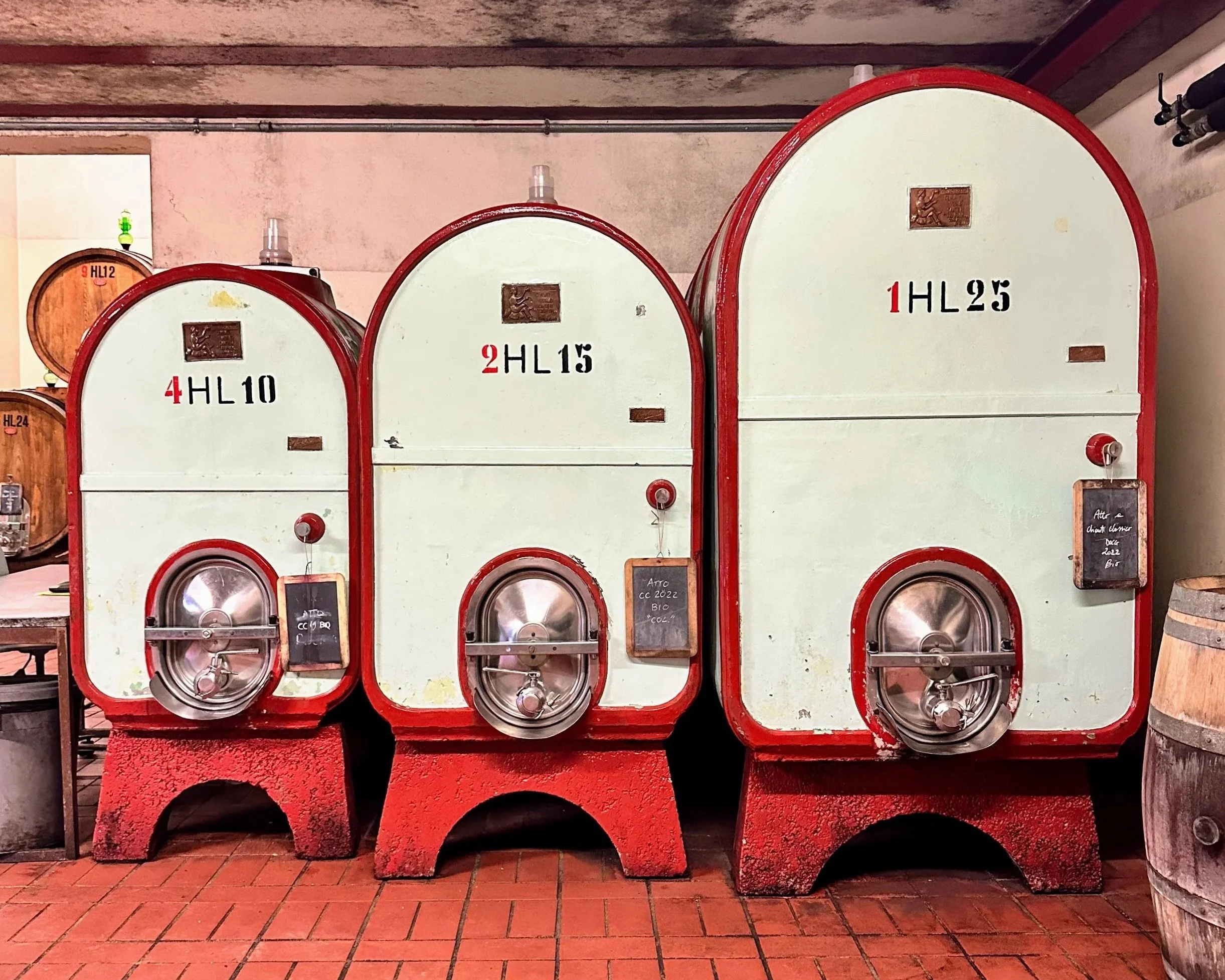 Three large vintage wine fermentation tanks with red borders, labeled 4HL10, 2HL15, and 1HL25, standing on red brick bases in a cellar.