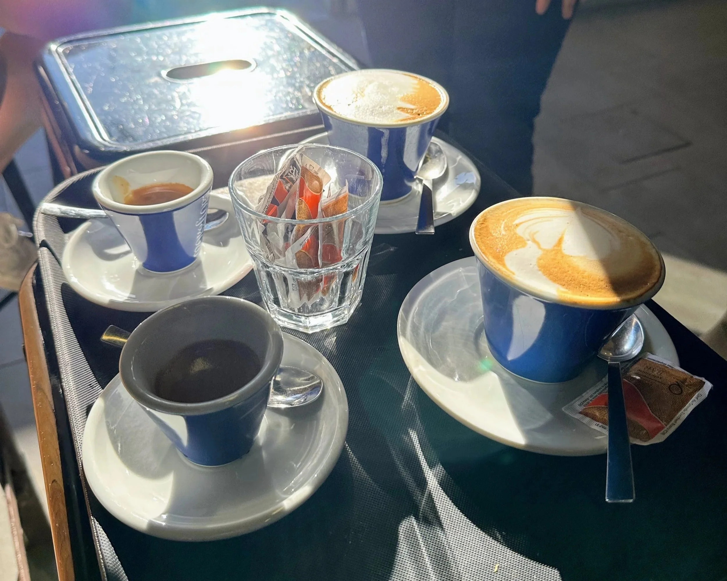 A tray with three cups of coffee or espresso, a glass of sweetener packets, and a small pot of cream or milk, on a black surface.