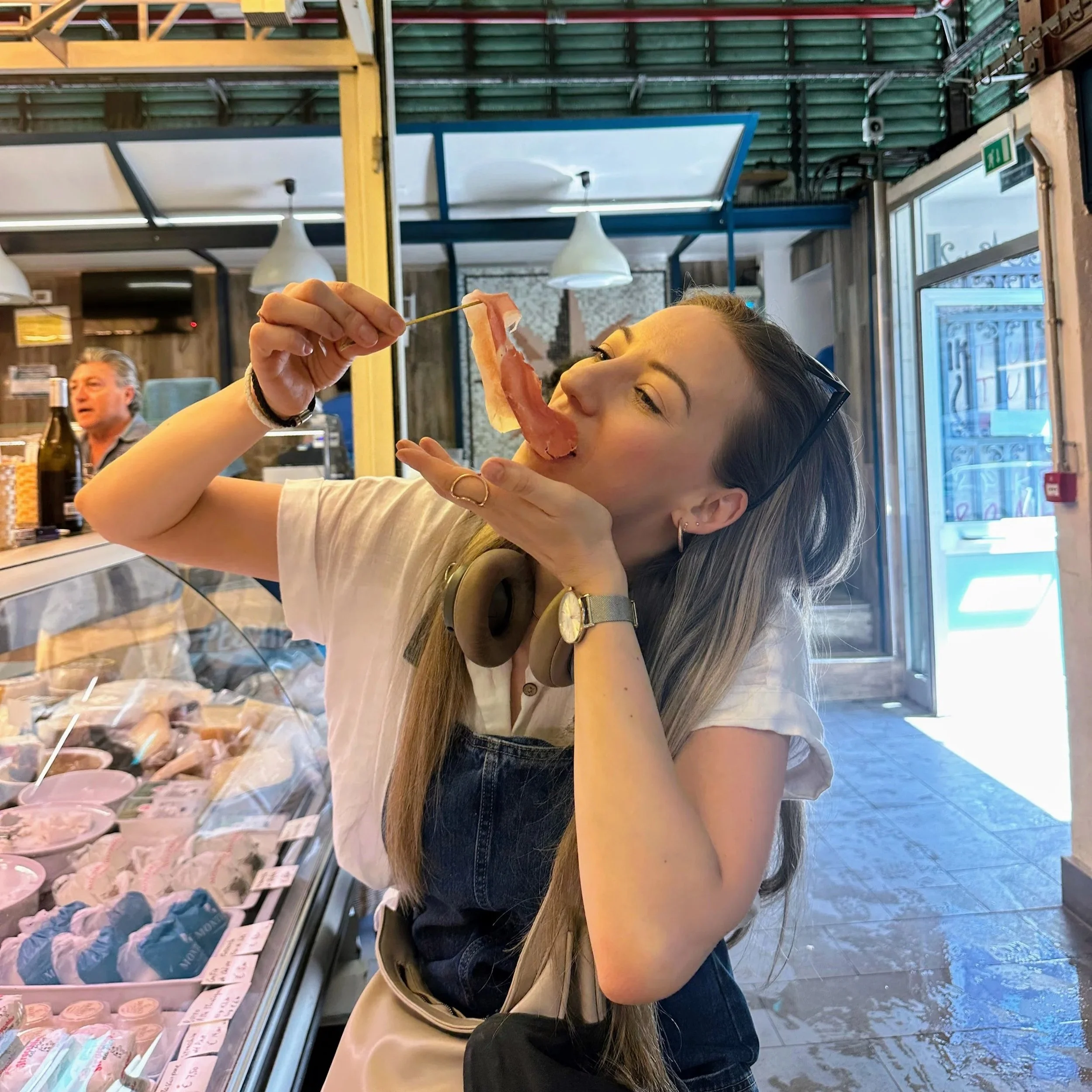 A woman with headphones around her neck and sunglasses on her head is holding a slice of prosciutto and pretending to eat it like a taco inside a delicatessen or food market.