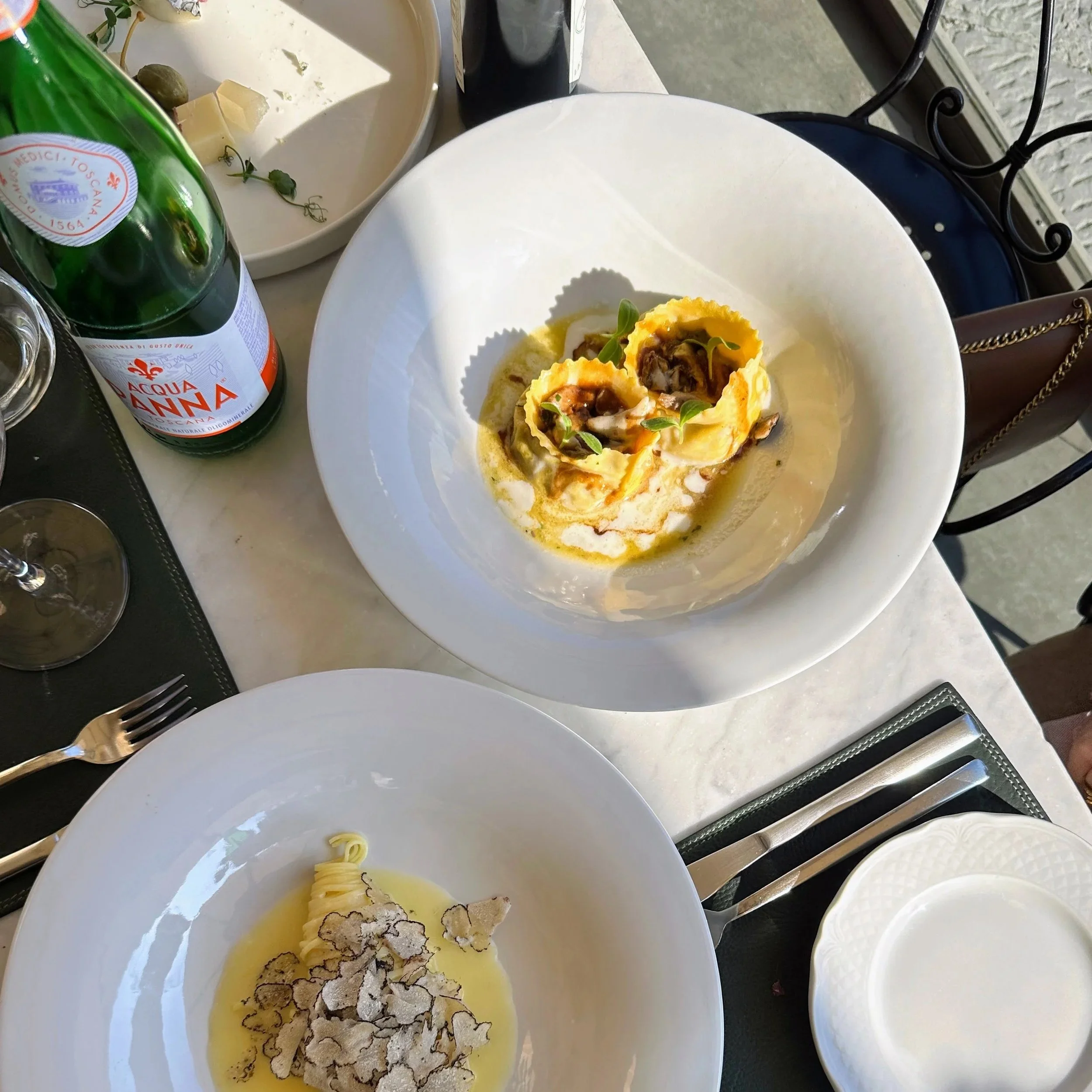 A table with two white plates of pasta, a bottle of sparkling water, a glass of water, a wine glass, and some utensils. One dish has ravioli with sauce and herbs, the other has pasta with truffle shavings and oil.