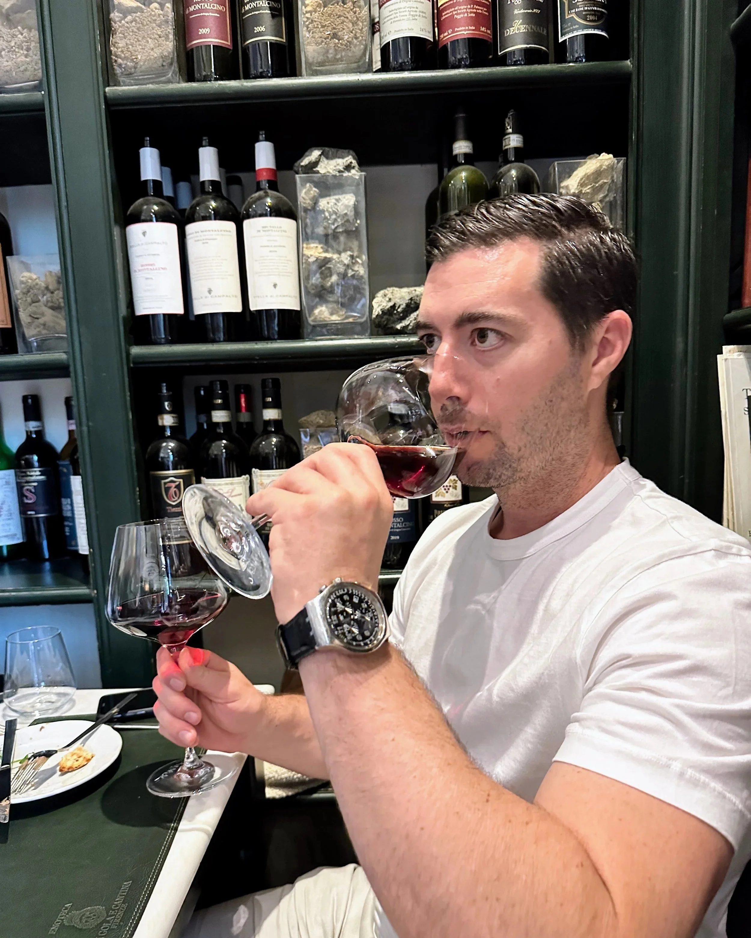 A man in a white t-shirt with a large watch on his left wrist is holding a glass of red wine and tasting it at a table in a restaurant or wine bar. Shelves with bottles of wine and rocks/ minerals are visible in the background.