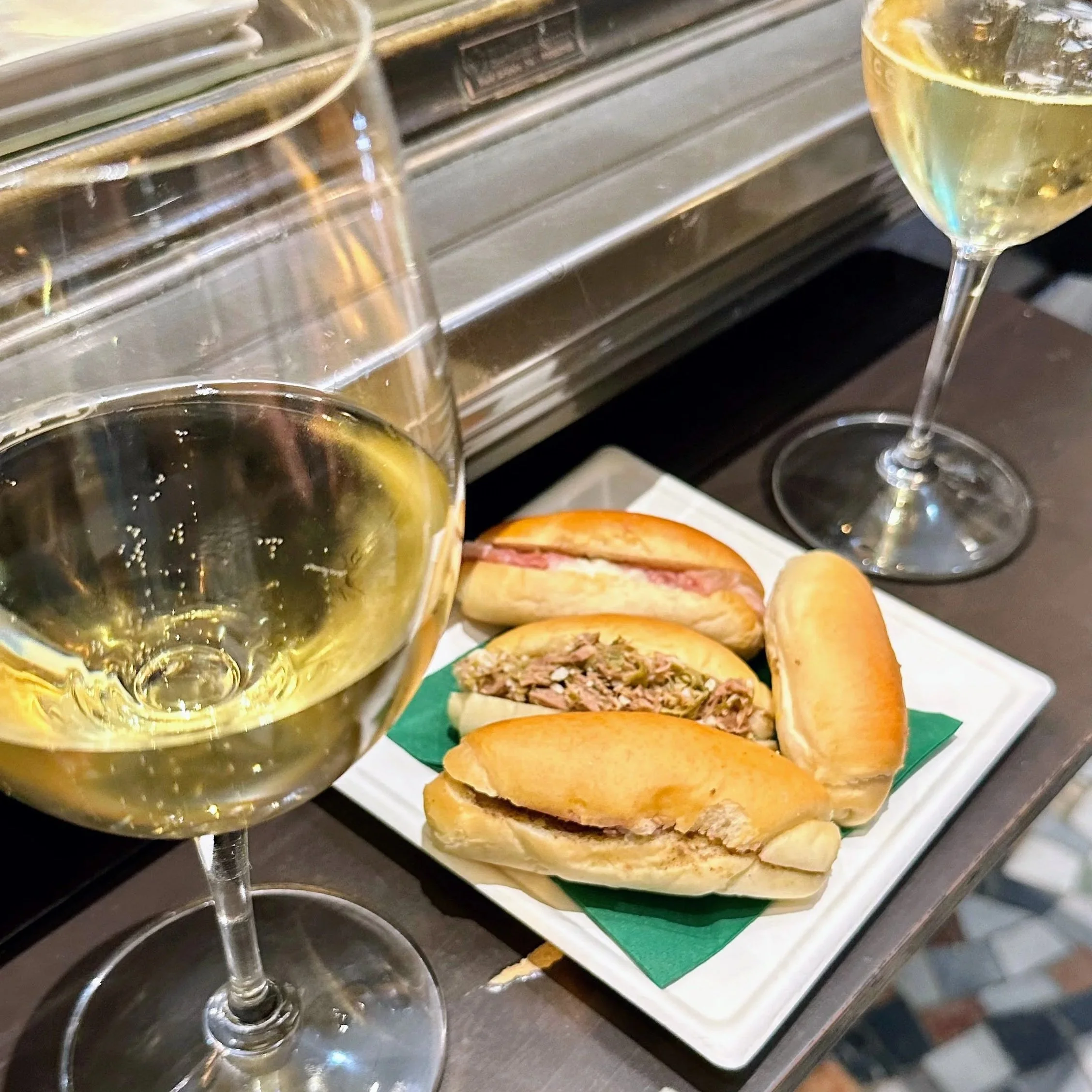 Two glasses of white wine and a plate with three mini sandwiches on a dark table.
