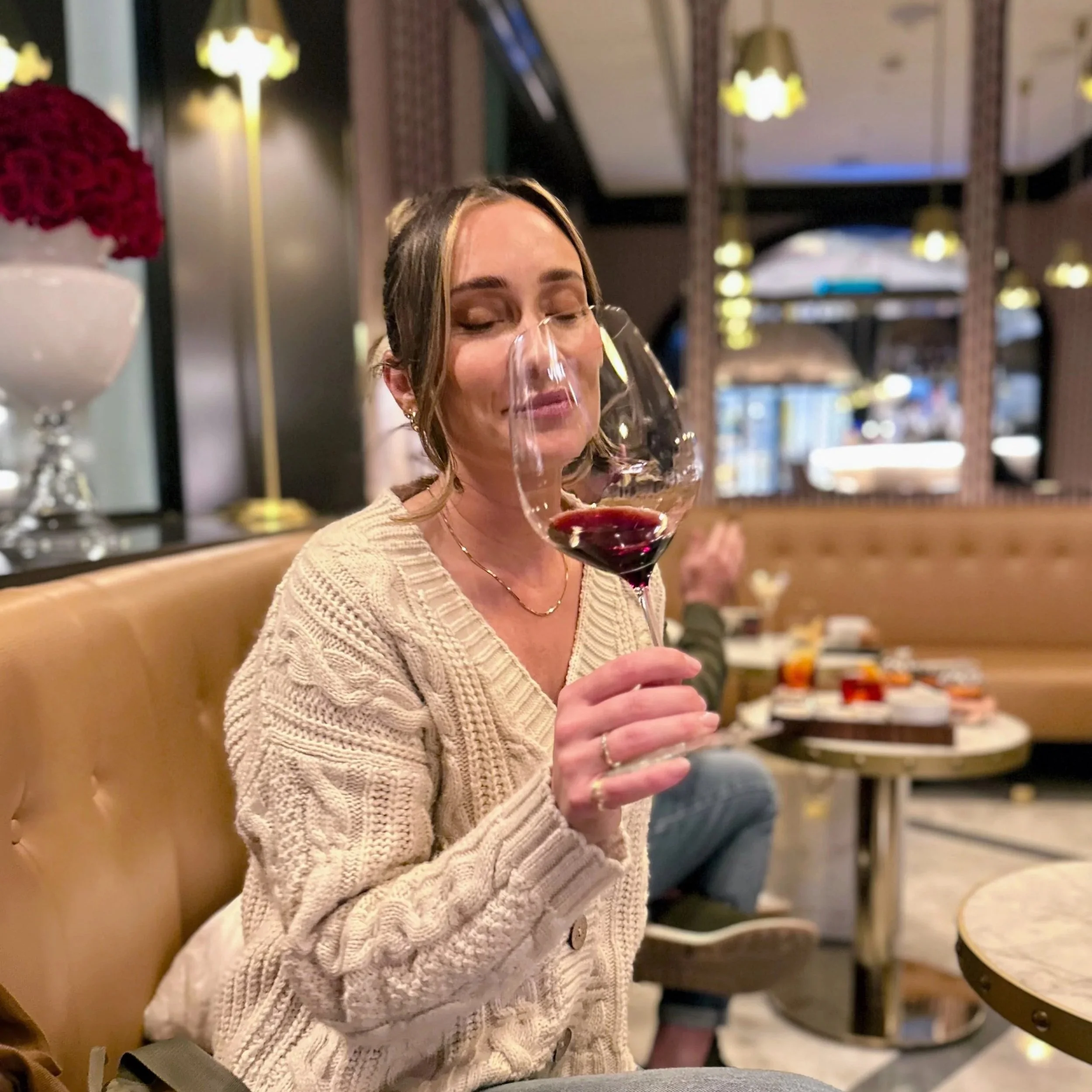 A woman sitting on a tan booth seat in a restaurant, holding a glass of red wine with her eyes closed, wearing a cream-colored knitted sweater.