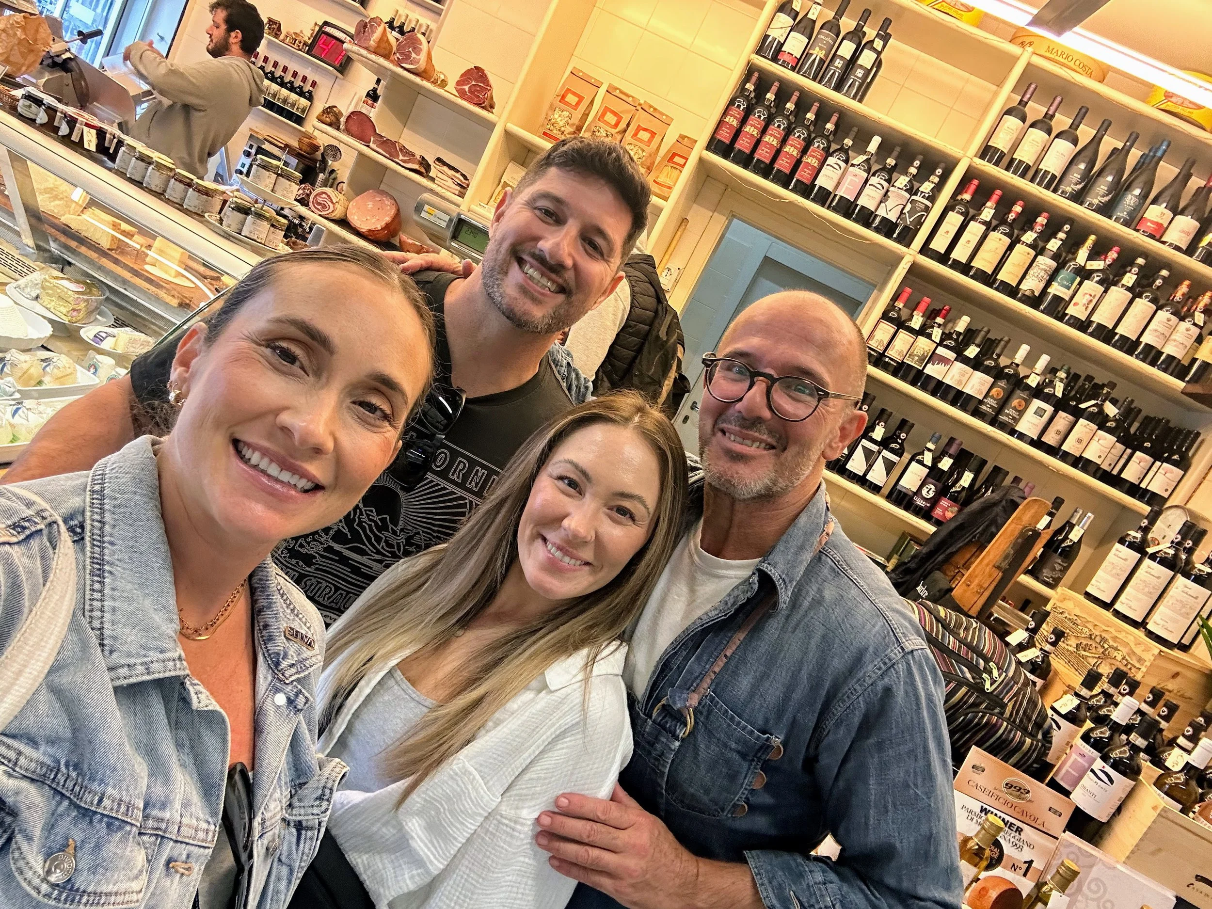 Four smiling people taking a selfie inside a wine shop or cellar, with shelves of wine bottles and a deli counter in the background.
