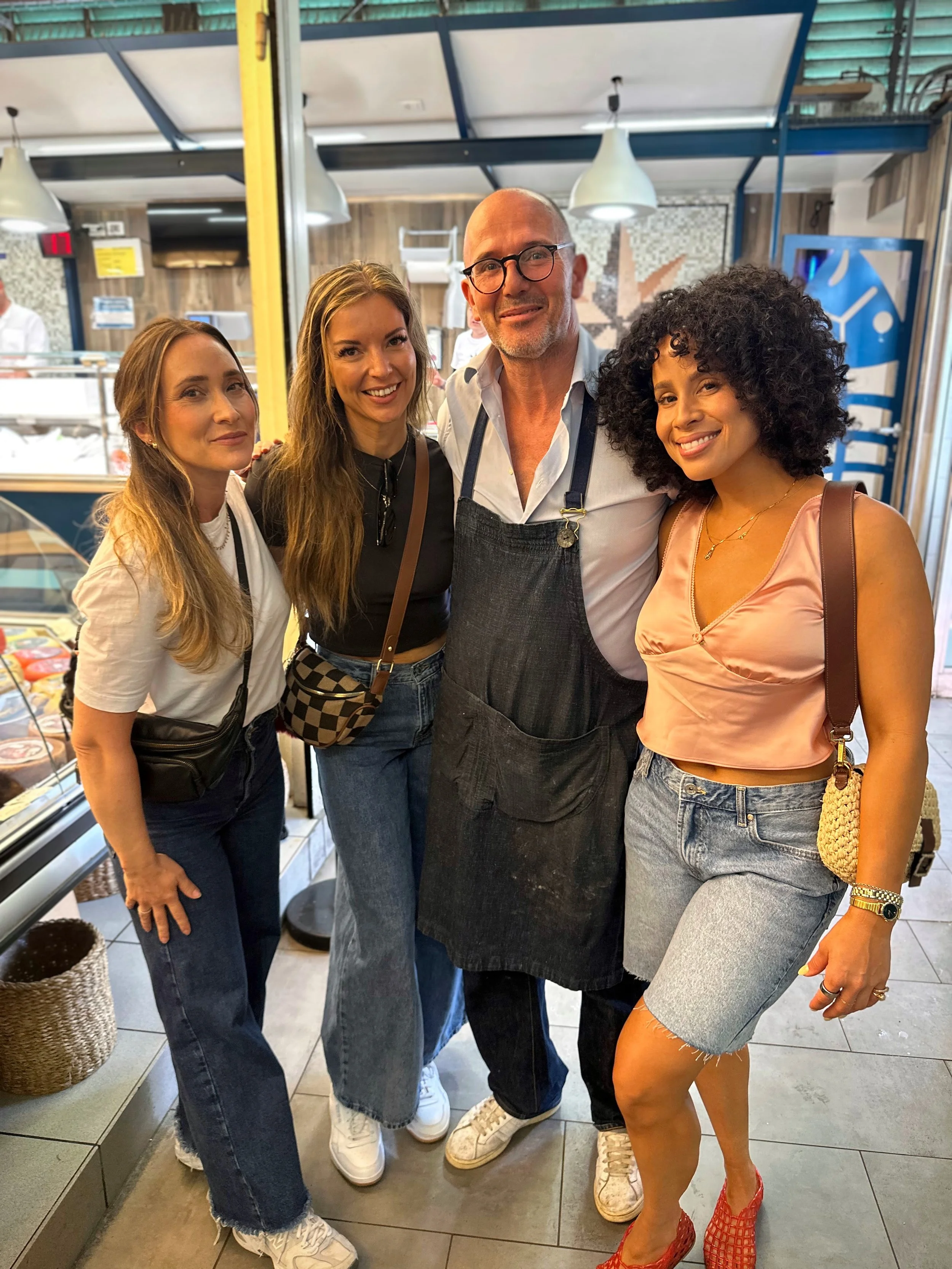 Four women and one man posing together inside a restaurant or deli, smiling at the camera. The man is wearing a black apron, while the women are dressed casually.