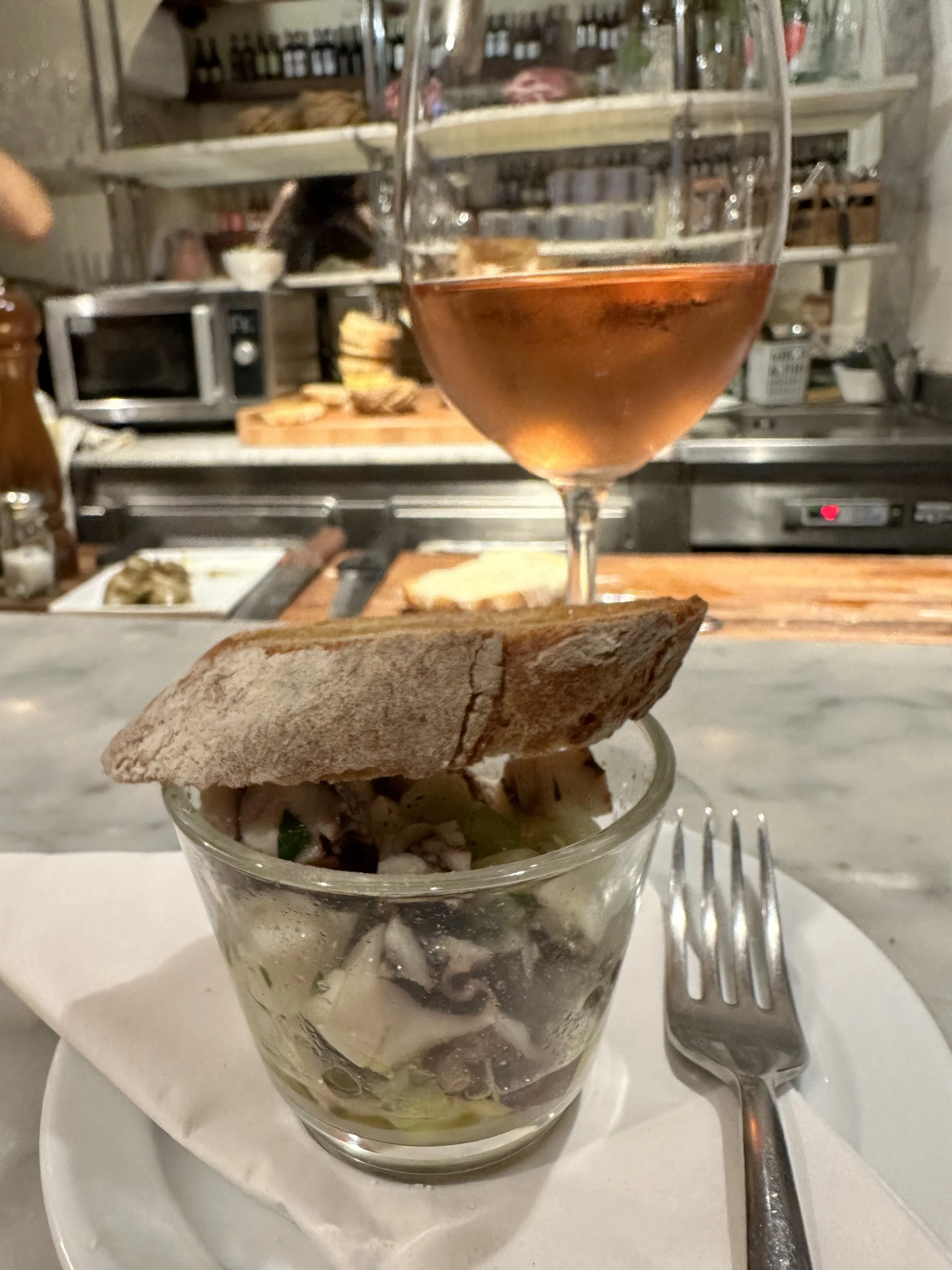 A glass of rosé wine placed on top of a small glass bowl filled with seafood salad, garnished with a slice of bread on top, on a white plate with a fork beside it.