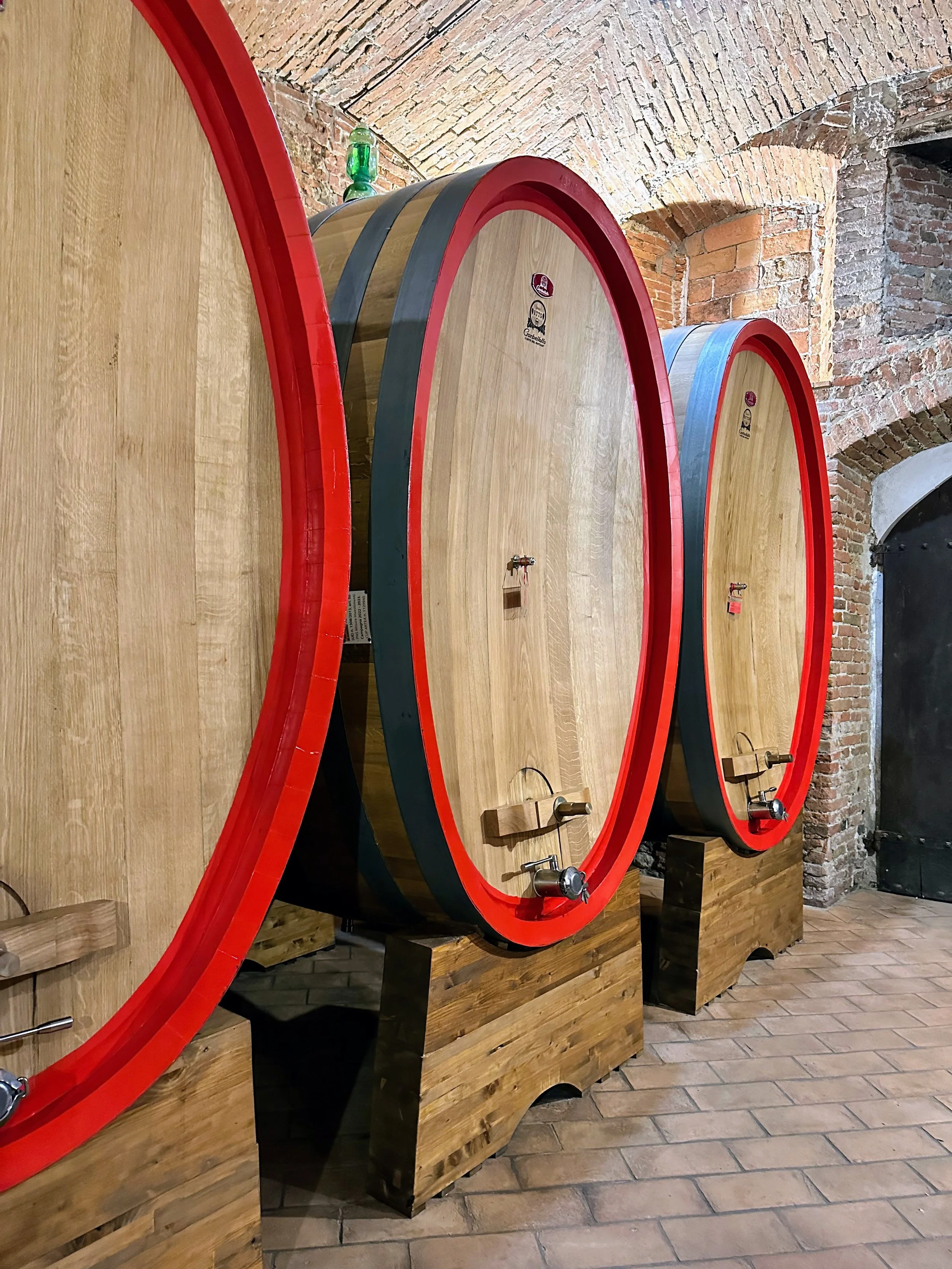 Three large wooden wine barrels with red rims and black bands in a cellar with brick walls and tiled floor.