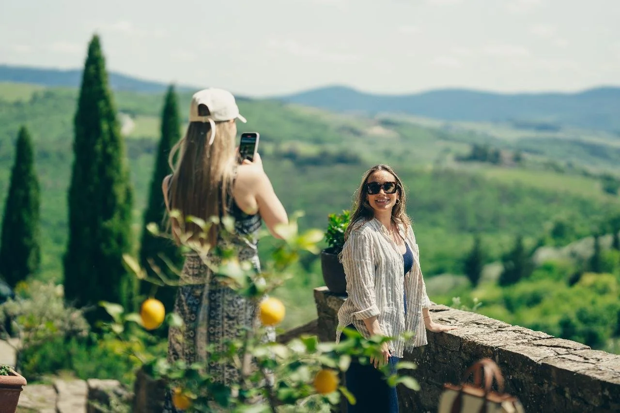 Two women outdoors in a scenic, hilly landscape with green foliage, one taking a photo of the other smiling in front of a stone wall.