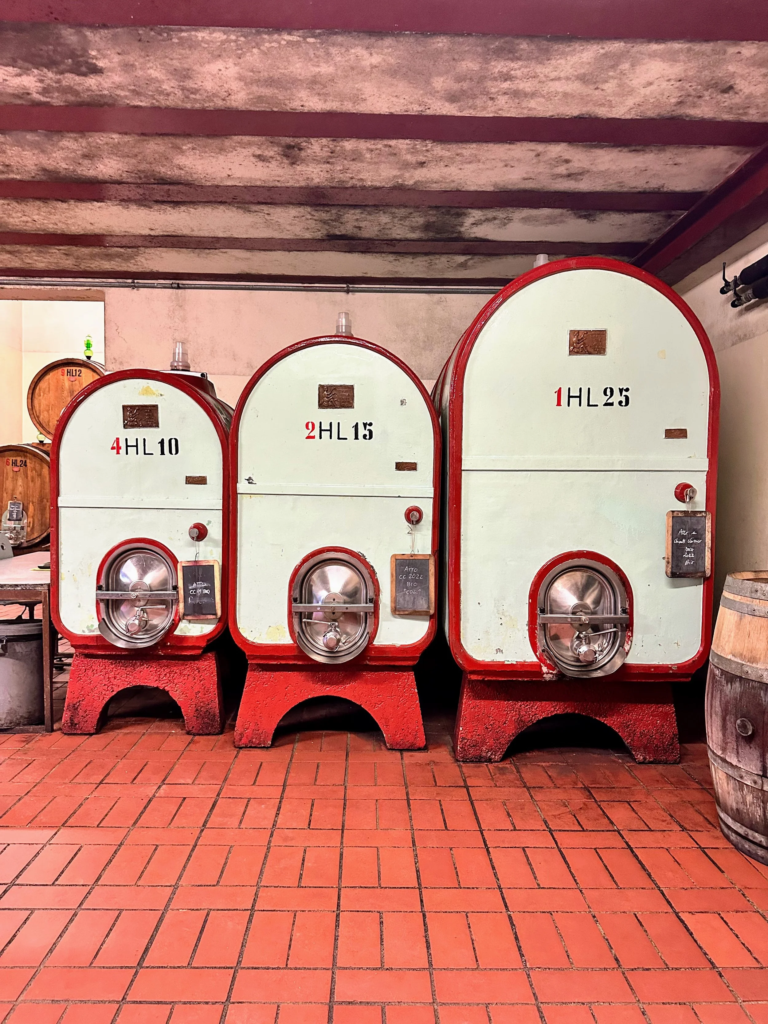 Three vintage wine fermentation tanks in a winery, labeled HL10, HL15, and HL25, standing side by side on red tiled floor with a rustic ceiling overhead.