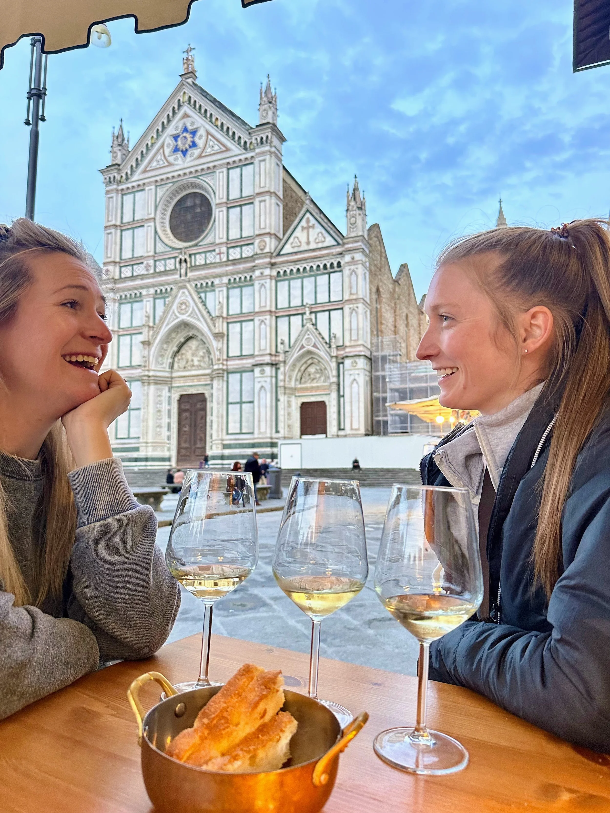A table set with two glasses of white wine, a bottle of water, a glass of water, two small bowls of snacks (green olives and round cookies), and an empty container on a brick surface.