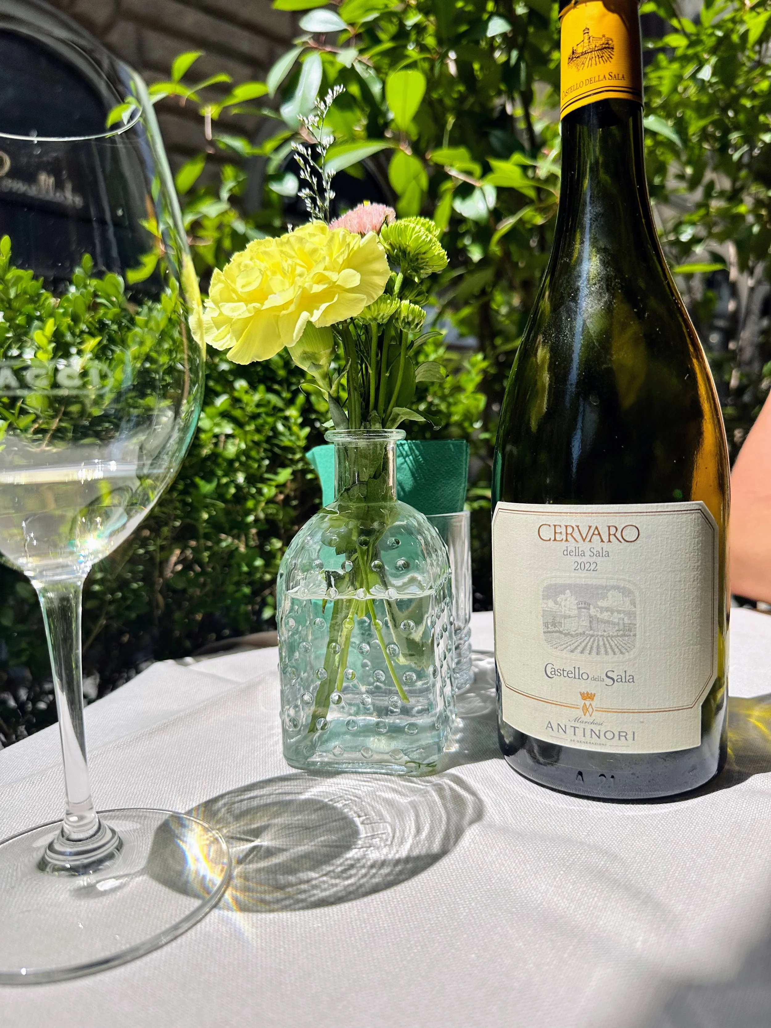A table setting with a glass of white wine, a small glass vase with yellow and green flowers, and a bottle of Cervaro della Sala 2022 wine on a white tablecloth, outdoors with greenery in the background.
