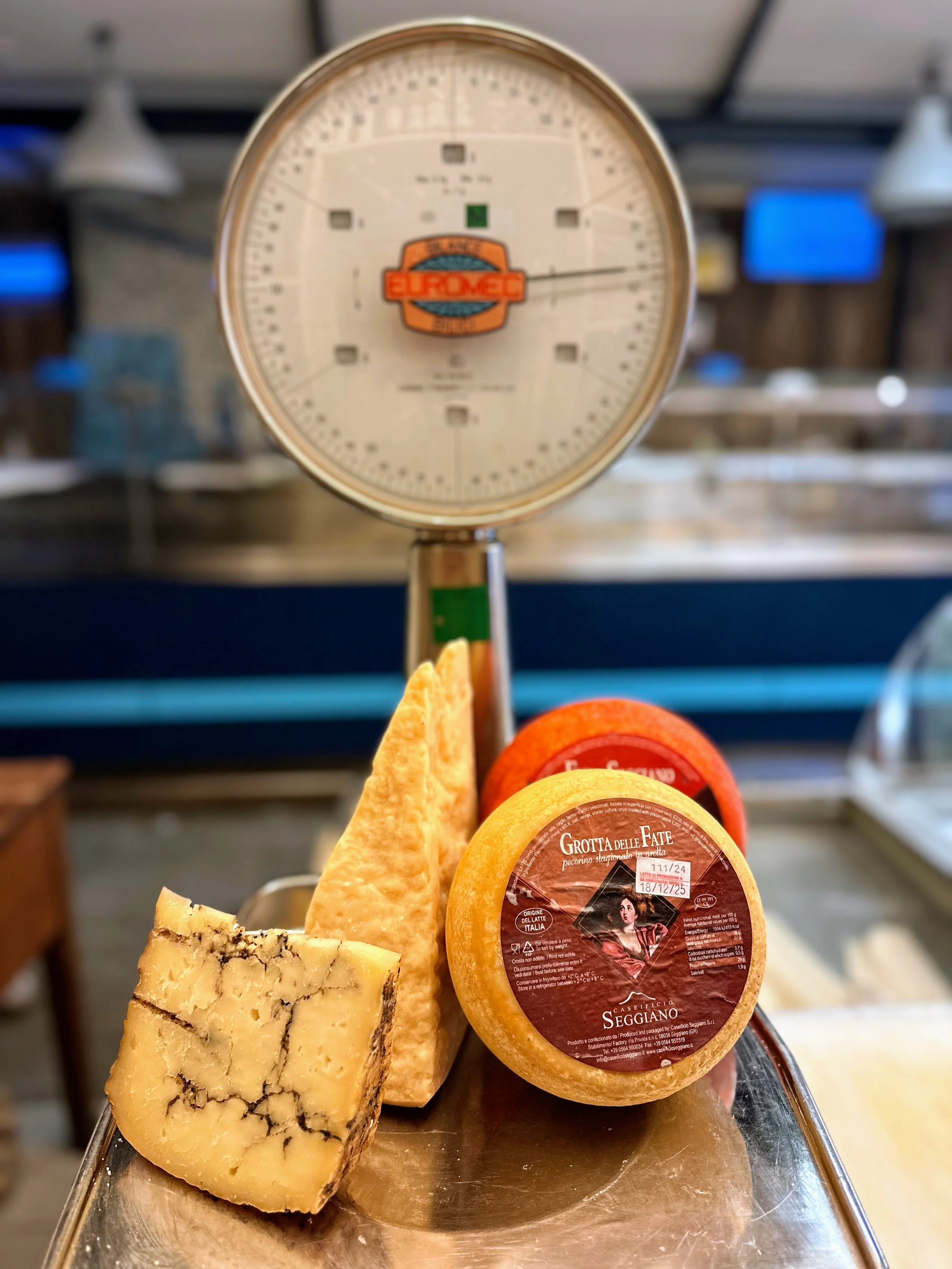 A cheese wheel labeled "Grotta Delle Fate" with slices of cheese, a wedge, and a vintage scale in the background.