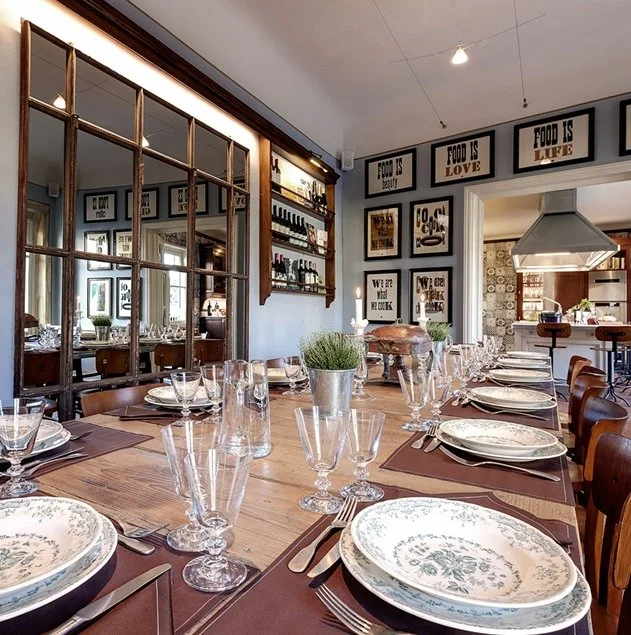 A dining room set for a meal with multiple glasses, plates, and silverware on a wooden table, decorated with a potted plant, and surrounded by framed wall art.