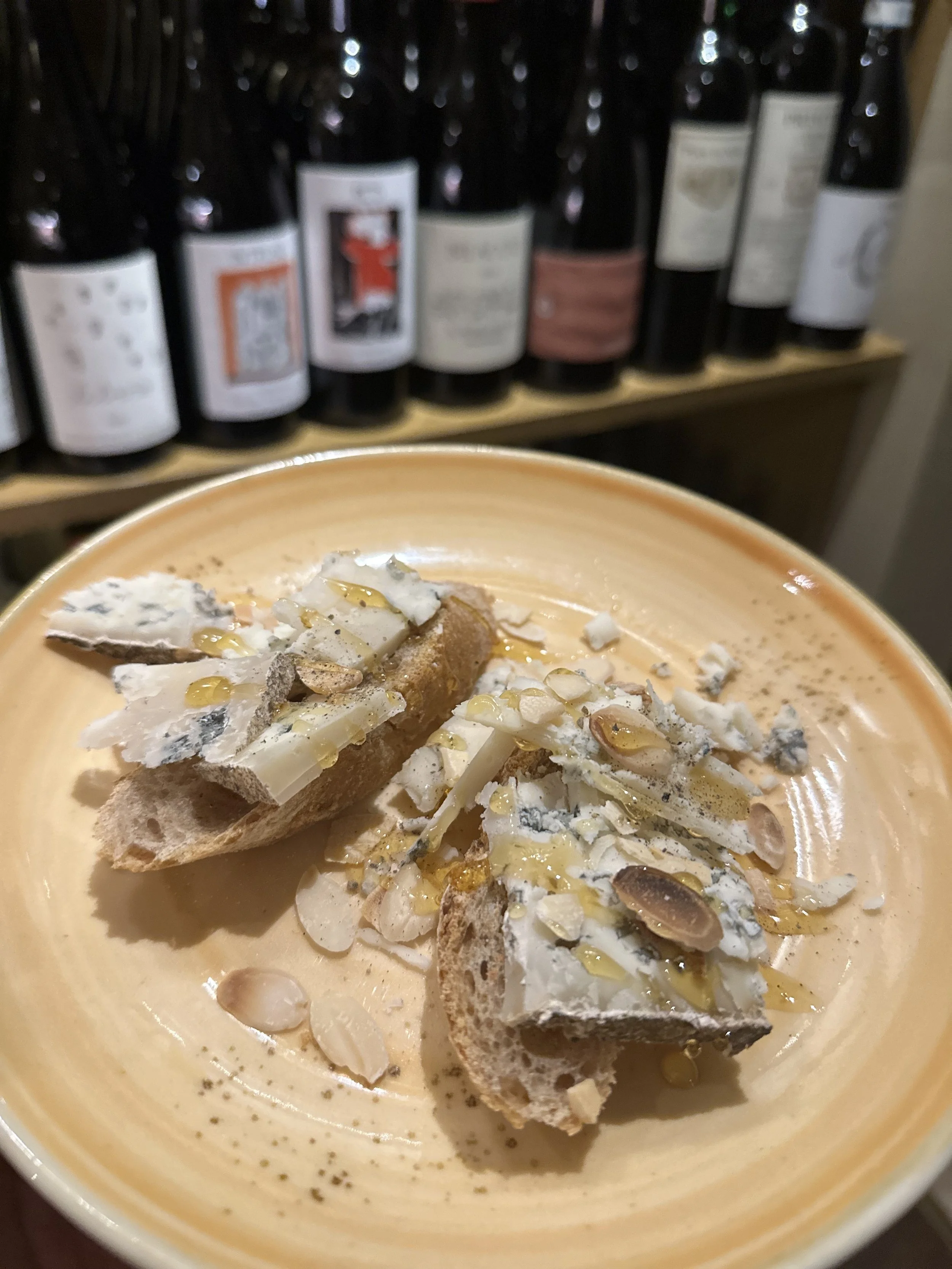 A bowl of blue cheese and toasted bread with blue cheese crumbles, sliced almonds, honey, and olive oil, in front of a wine rack.