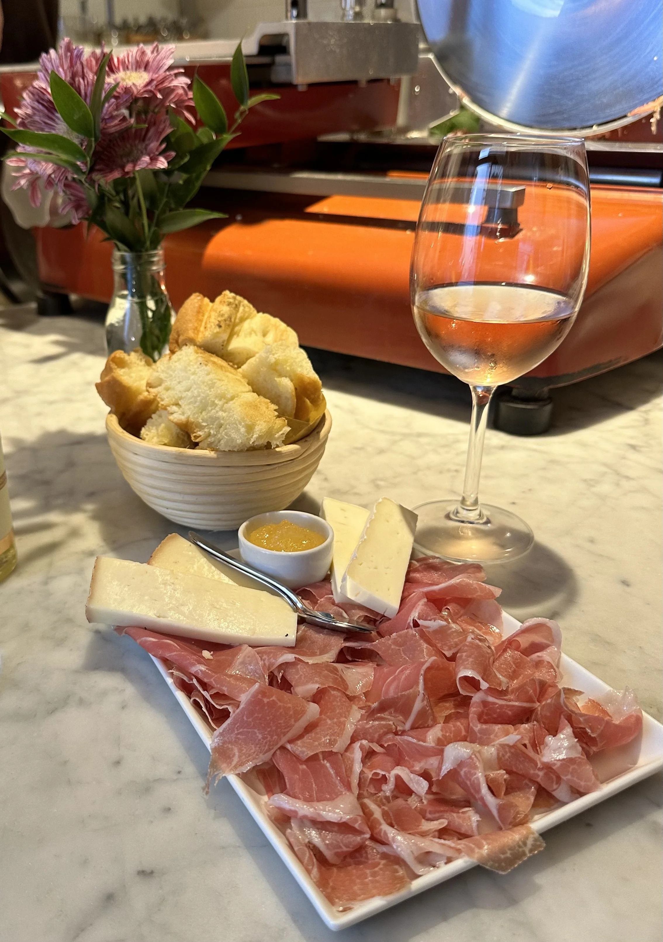 Charcuterie board with sliced prosciutto, cheese, and mustard, along with a glass of white wine, bread chunks in a bowl, and a small vase of purple flowers on a marble countertop.