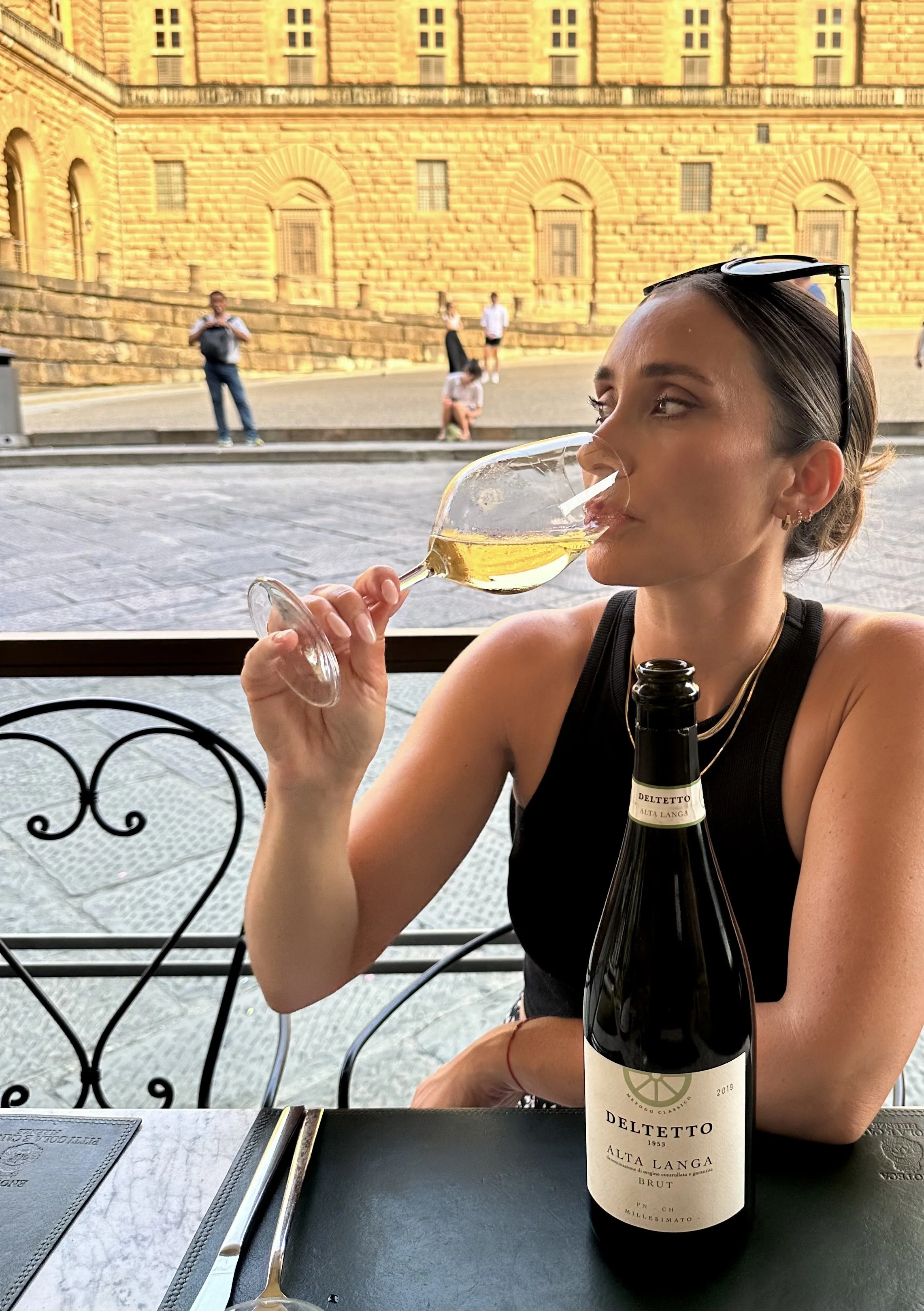 A woman with sunglasses on her head is sitting at a table indoors, drinking white wine from a glass. There is a bottle of Deletto Alta Langa Brut wine on the table. Through the window behind her, a historic building and several people walking outside