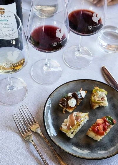 Three glasses of red wine, one glass of white wine, and a plate with four small appetizers on a white tablecloth.