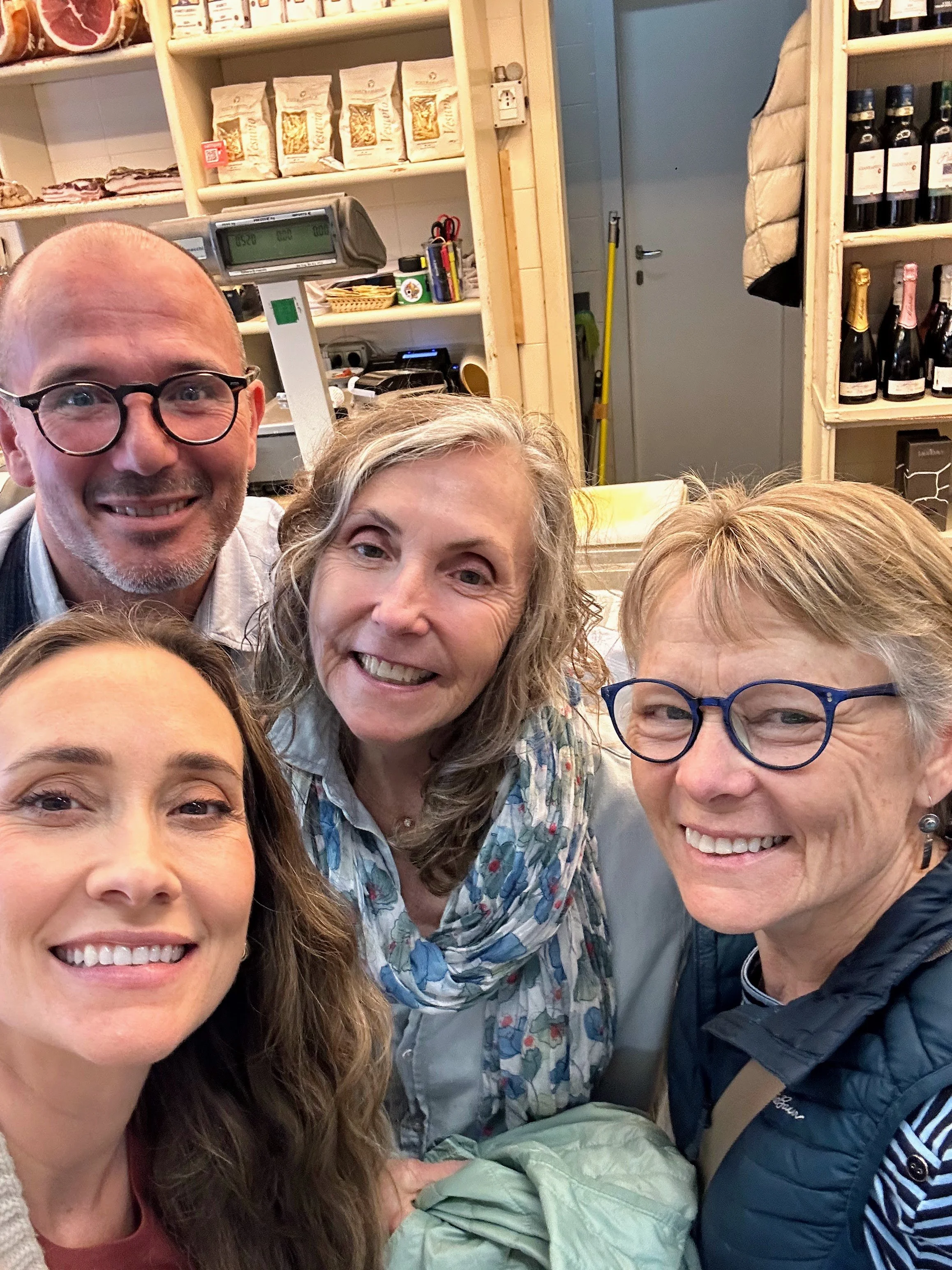 Four smiling women and one smiling man taking a selfie inside a store or cafe, with shelves of food and drinks in the background.