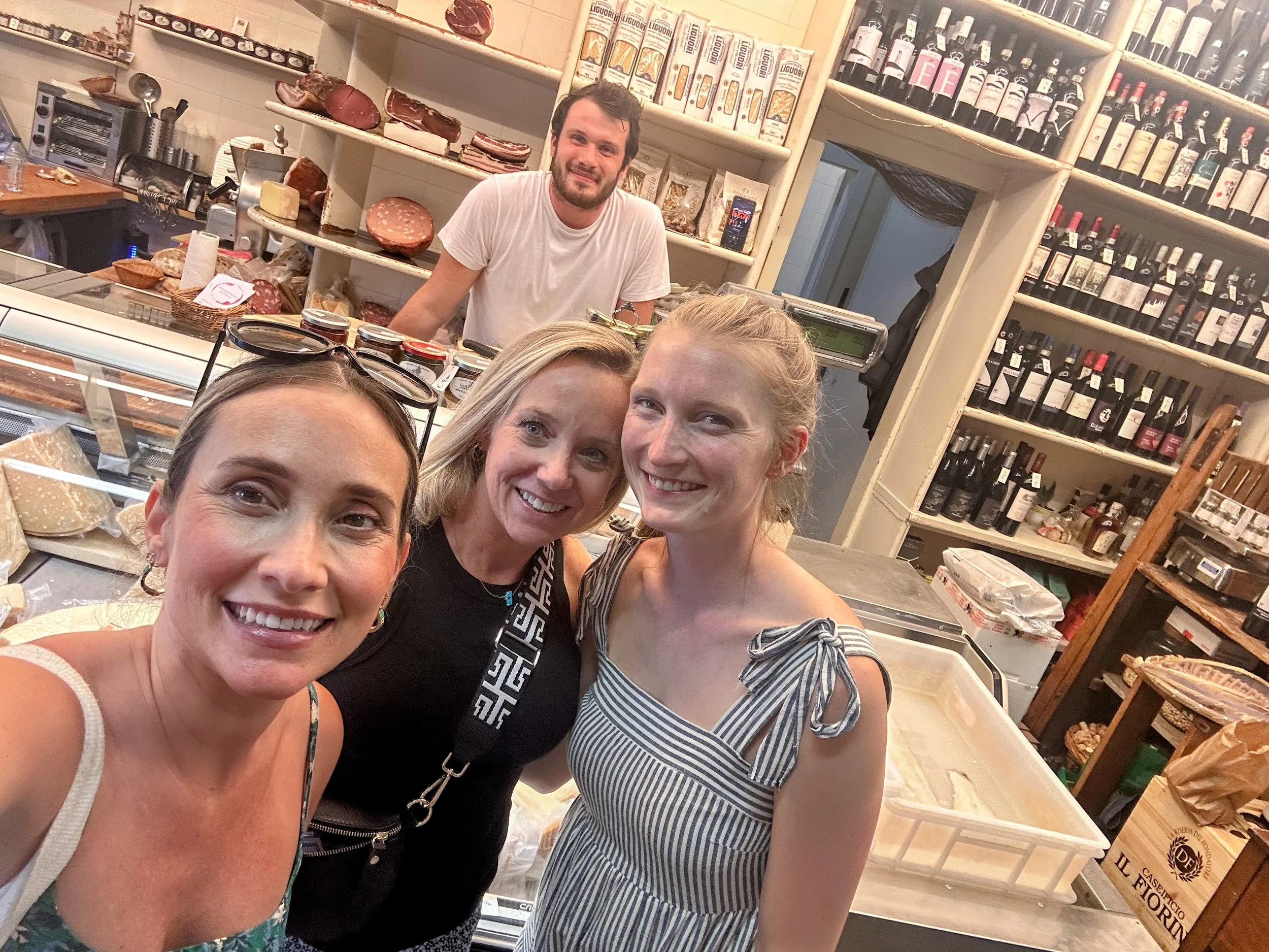 Four smiling women taking a selfie inside a cheese and wine shop, with shelves of wine bottles and cheese behind them, and a man working at the counter in the background.