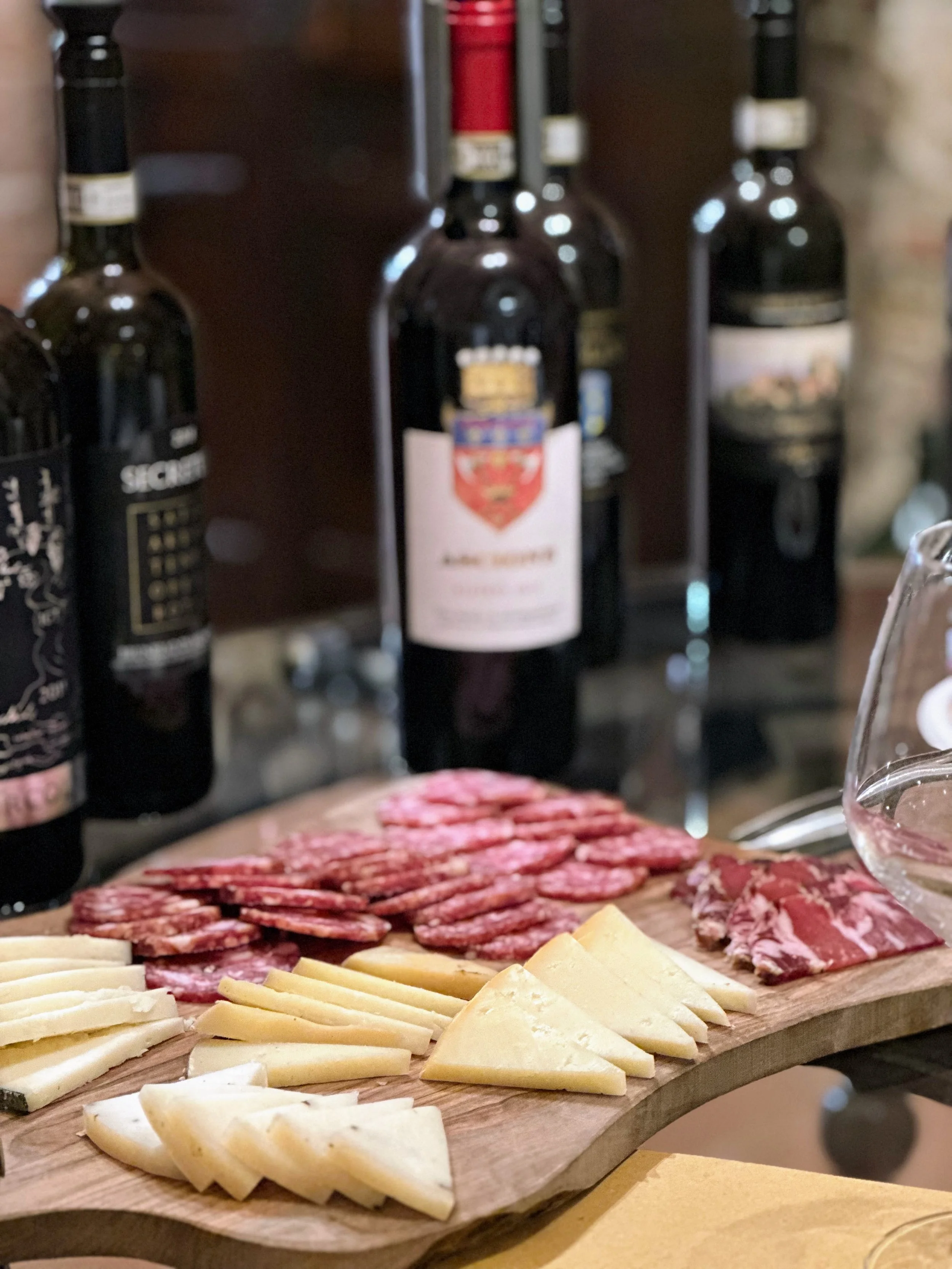 A wooden charcuterie board with sliced cheeses and assorted cured meats, set in front of a blurred background of wine bottles.