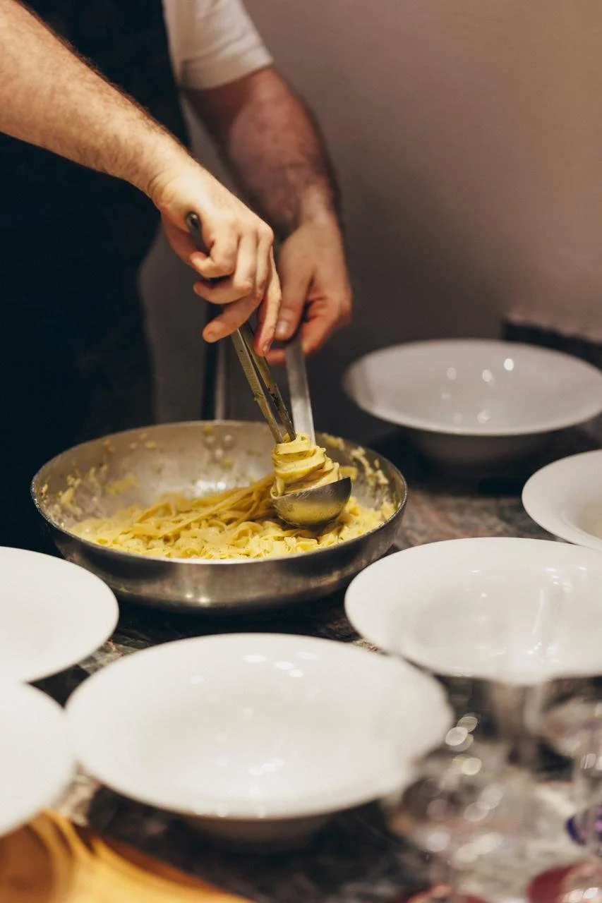Person serving spaghetti pasta onto a plate in a dining setting with empty bowls around.