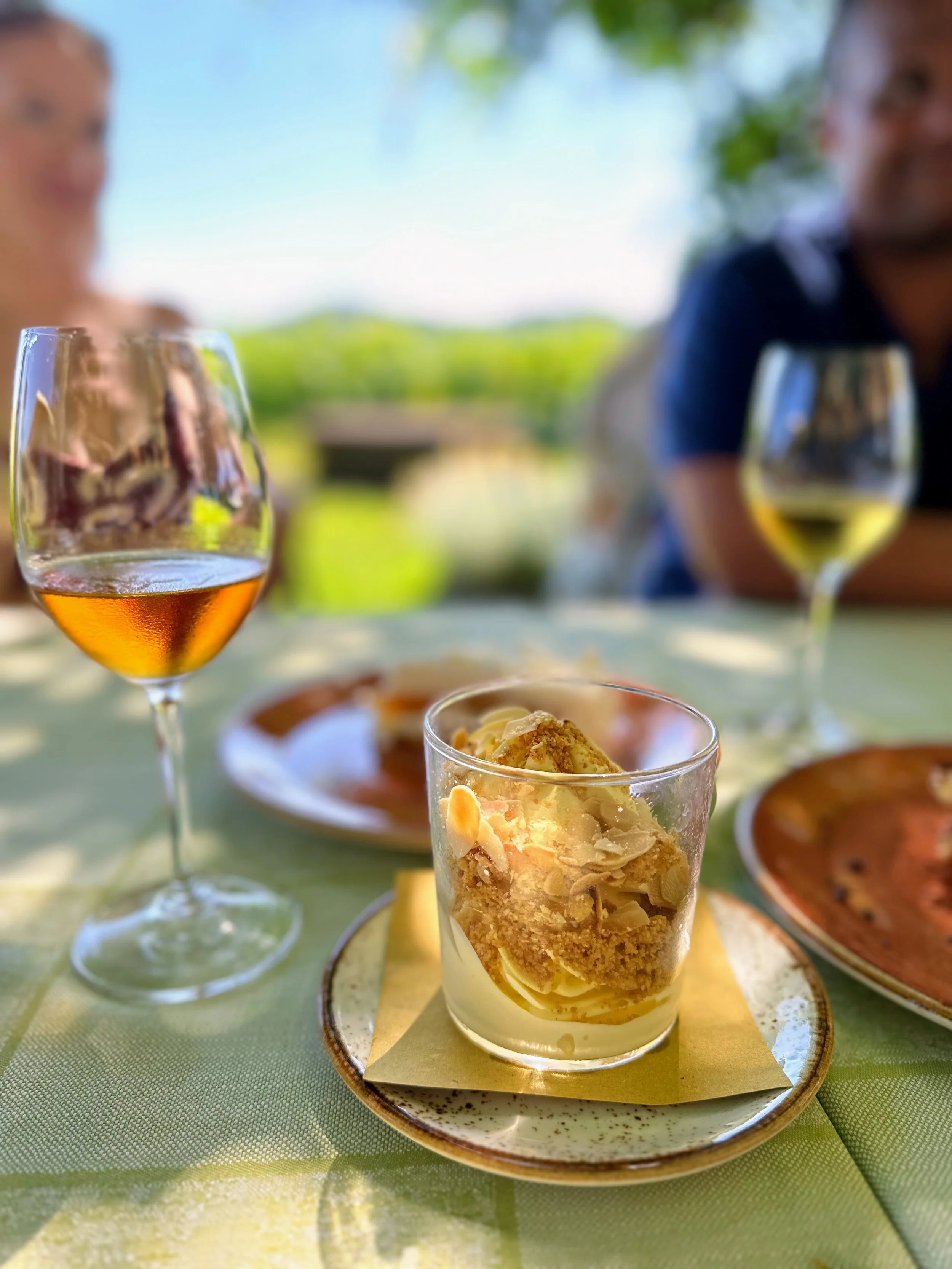 Dessert in a glass cup topped with whipped cream and nuts, with two glasses of white wine and plates of food on a table outdoors, with people in the background.