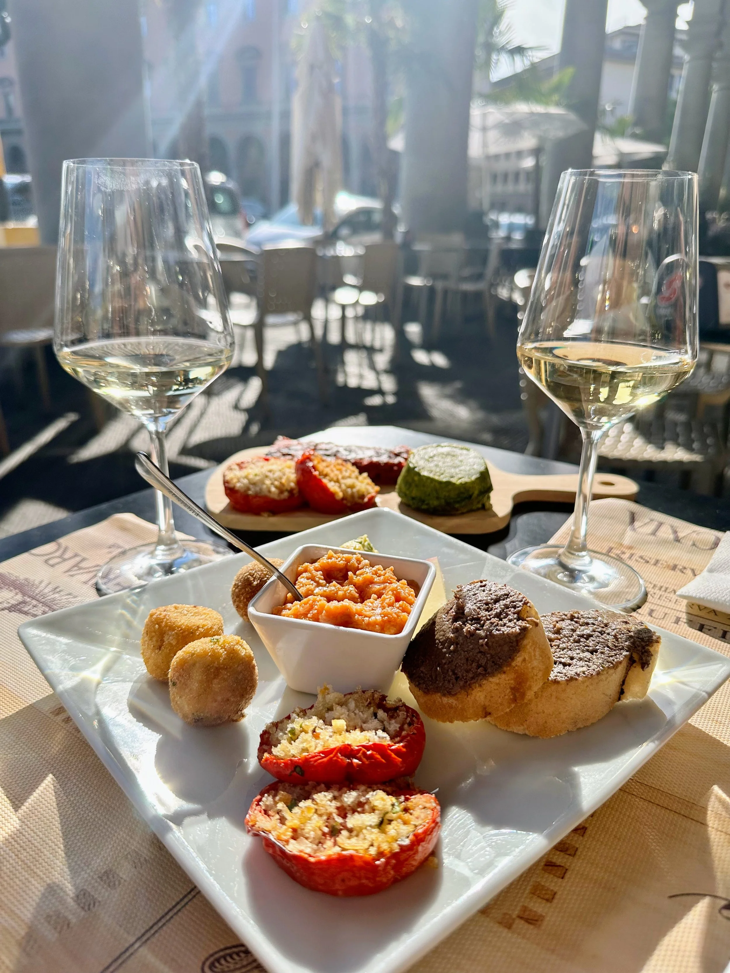 A white plate with various Italian appetizers including stuffed tomatoes, croquettes, breaded eggplant, and a small cup of tomato sauce, with two glasses of white wine, set on a table near a window with sunlight streaming in.
