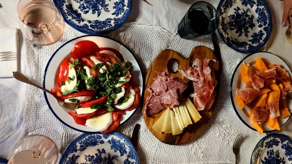 A dinner table with a variety of foods including a caprese salad with tomatoes, mozzarella, and basil, a meat and cheese platter with prosciutto and other cured meats, a bowl of sliced cantaloupe or similar fruit with prosciutto, and glasses of rosé