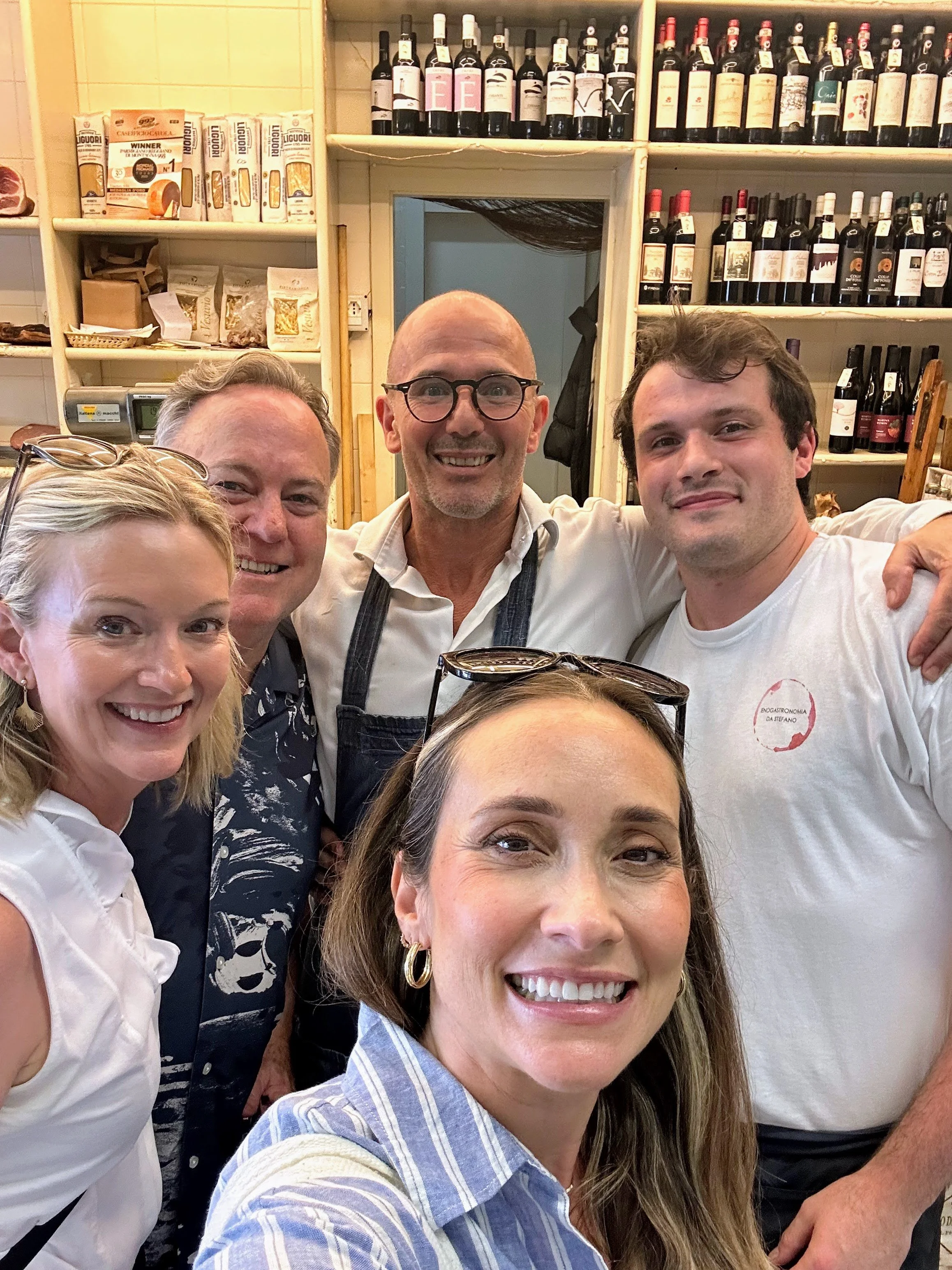 Group of five people smiling and taking a selfie in a wine shop or restaurant, with shelves of wine bottles and food products in the background.