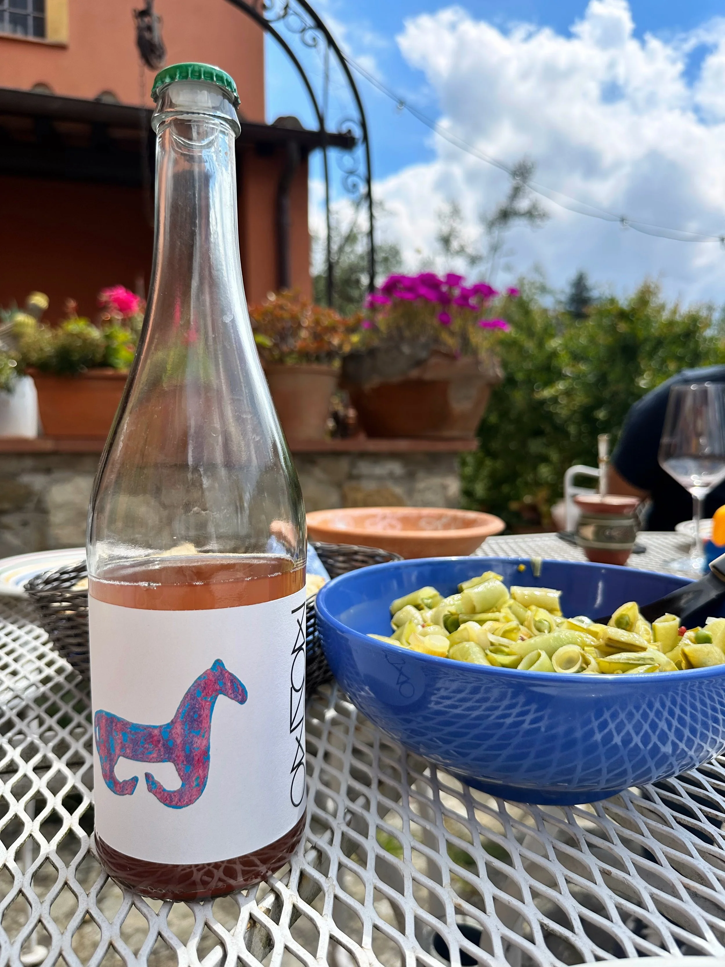 A glass bottle with a colorful dog illustration next to a blue bowl filled with cooked green beans on a white mesh table outdoors, with potted plants and a partly cloudy sky in the background.