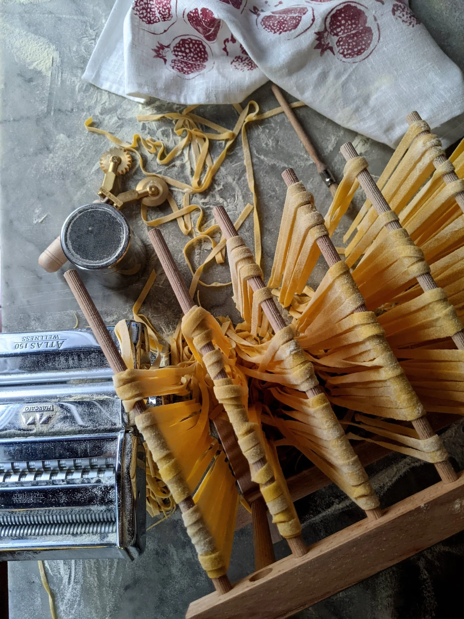 Uncooked pasta cones with wooden sticks set on a pasta drying rack, surrounded by a pasta maker, a jar of flour, and a towel with a strawberry print.