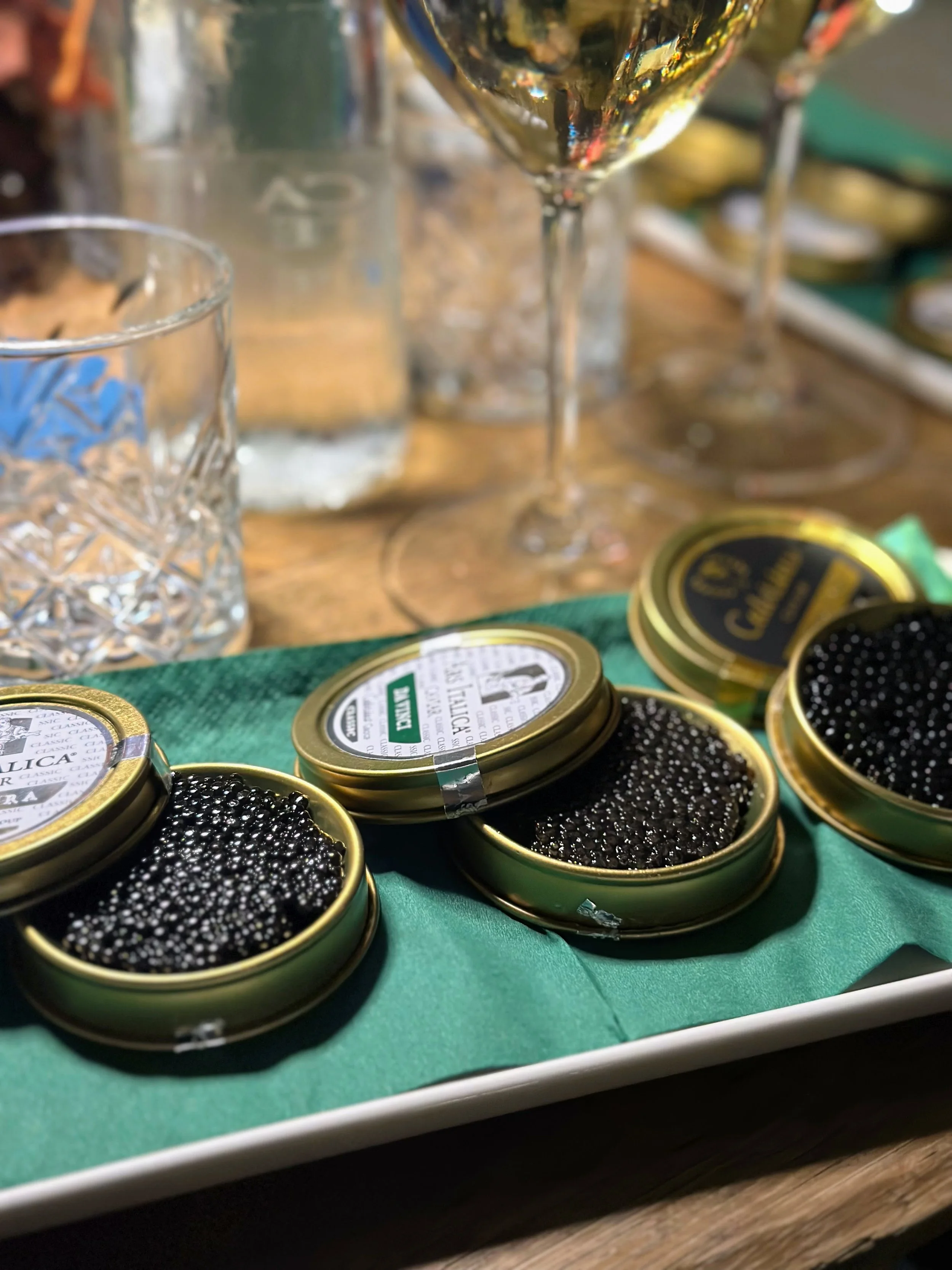 Open tins of black caviar on a green napkin with glasses and bottles in the background.
