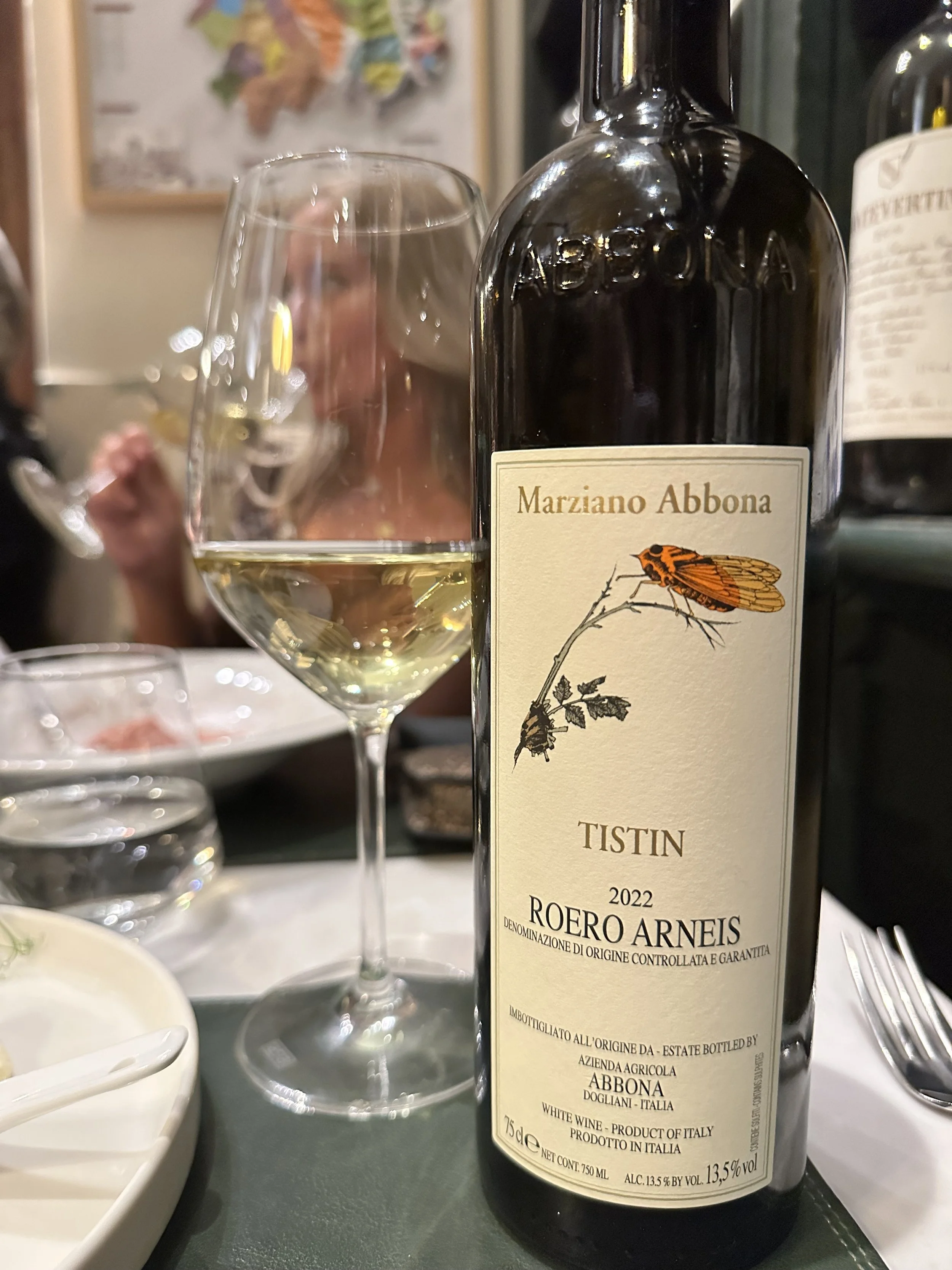 A bottle of Marziano Abbona Roero Arneis white wine next to a filled wine glass on a table, with a blurred woman in the background eating in a restaurant.