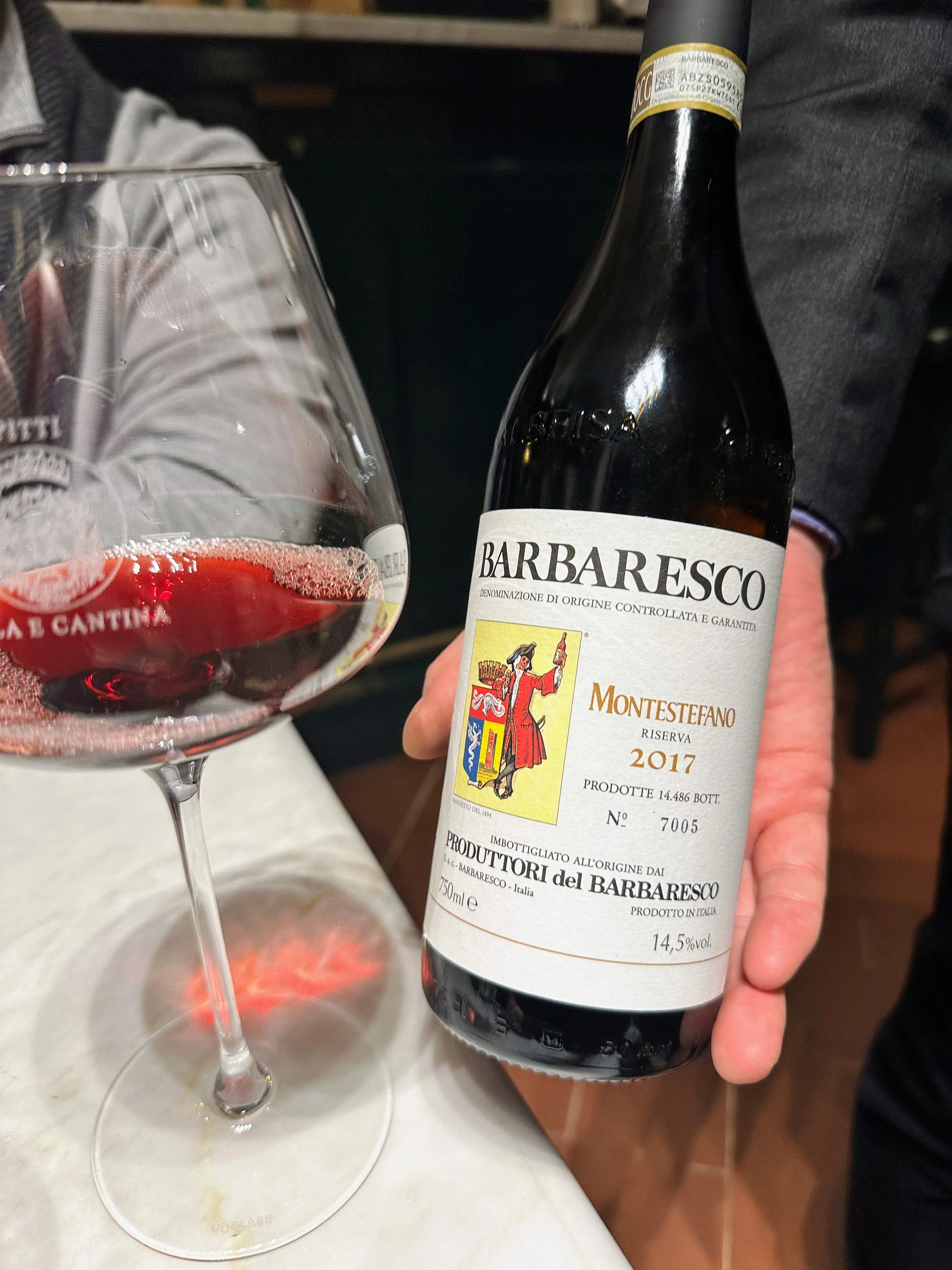 A person holding a bottle of 2017 Barbaresco Montesteffano red wine next to a filled wine glass on a white surface.