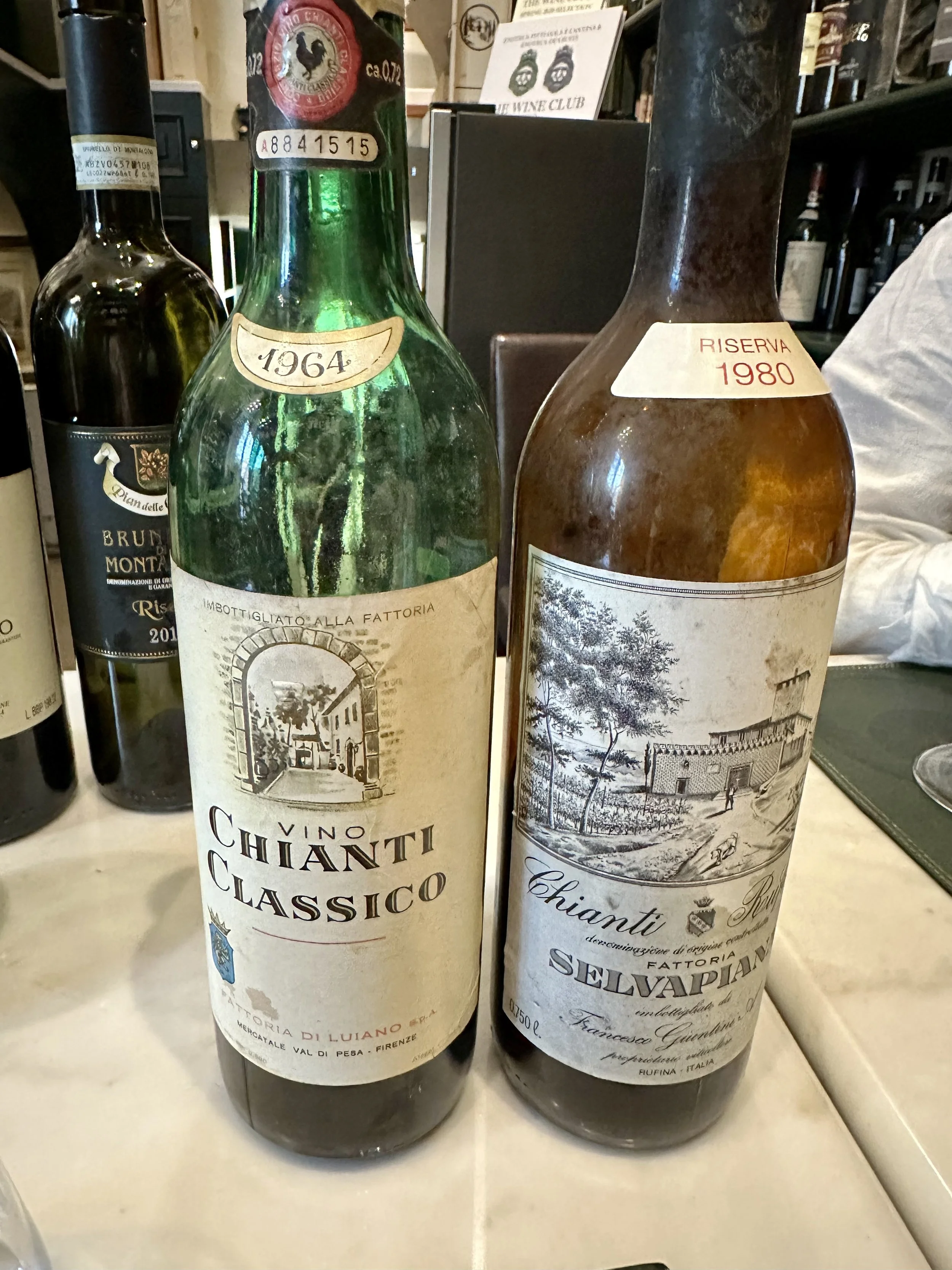Two bottles of Chianti wine placed on a table, one labeled Chianti Classico from 1964 and the other labeled Chianti Riserva from 1980.